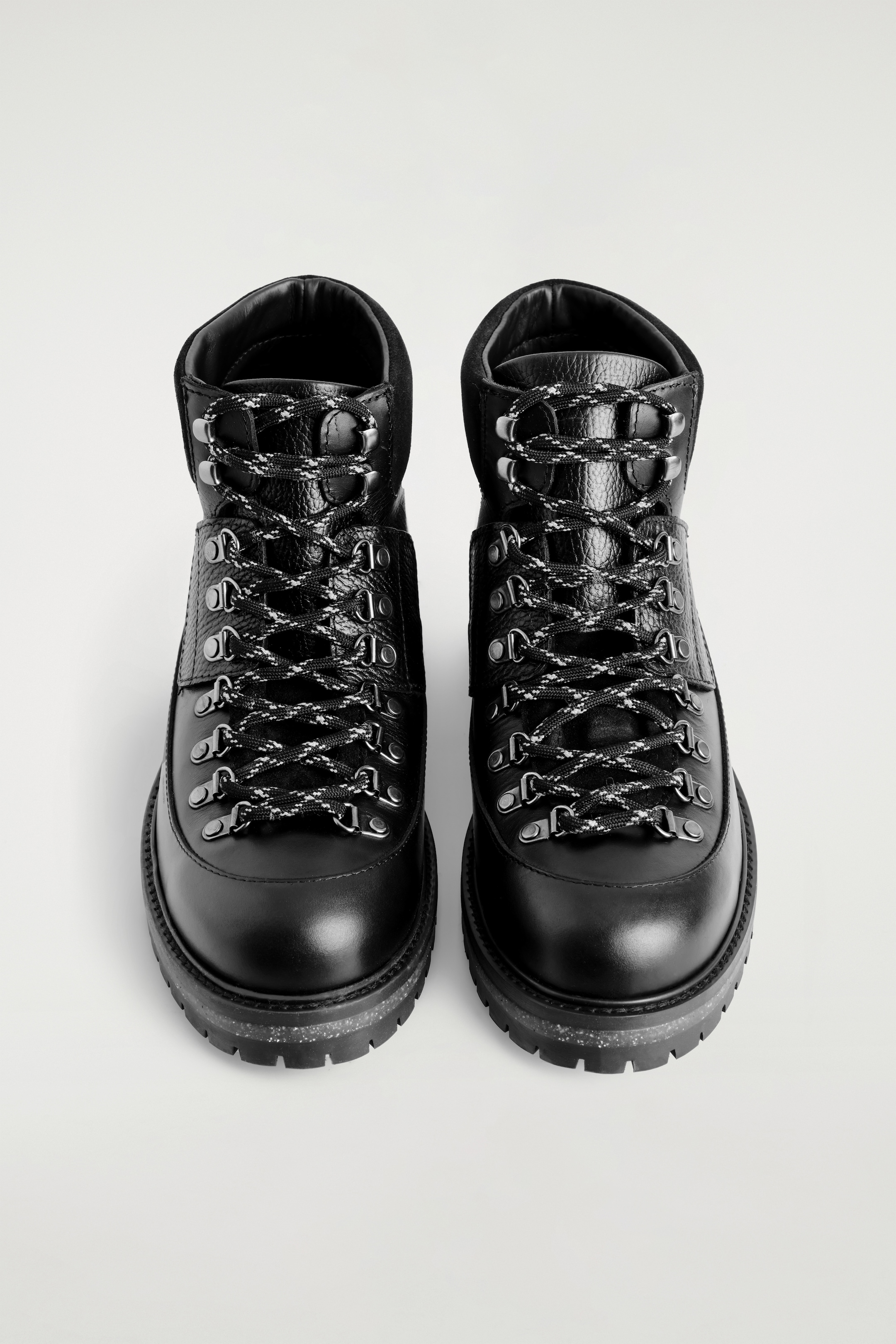 Agrandir l'image: Deux bottes noires inspirées de la randonnée sont placées côte à côte, face à l'avant, mettant en valeur leur construction en cuir lisse et grainé avec des colliers de cheville en daim rembourrés. Elles sont dotées de lacets en corde noirs et blancs épais, enfilés à travers des anneaux en D argentés et des crochets rapides, et reposent sur des semelles épaisses et superposées avec des sections médianes mouchetées.