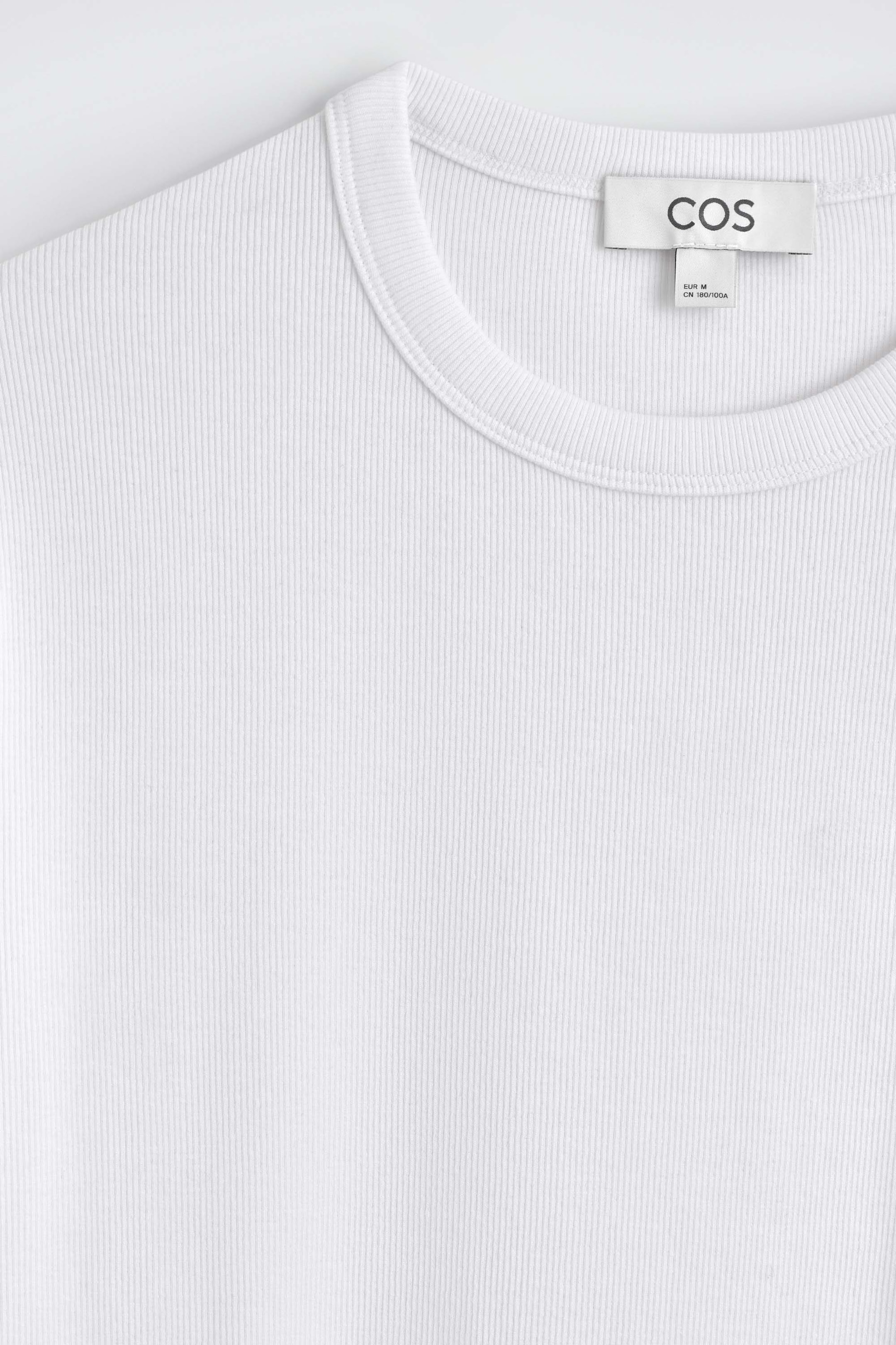 The white slim-fit ribbed cotton T-shirt features a ribbed crew neck and a fine vertical ribbed texture in a close-up front view. A white brand label is stitched inside the back collar of the garment.