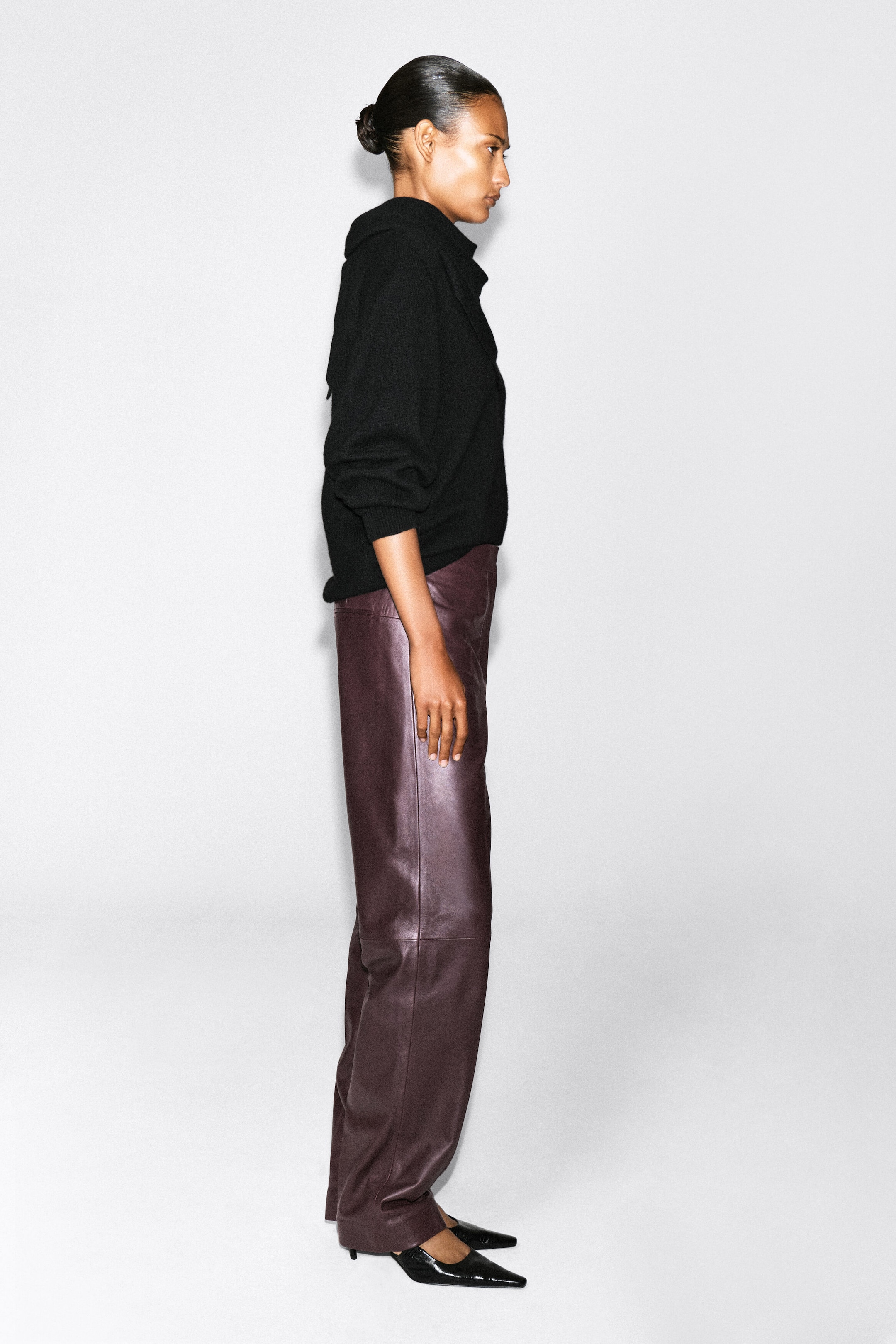 The model is standing in profile wearing burgundy leather straight-leg pants. The pants feature a smooth texture and a relaxed fit, styled with a black long-sleeved sweater and black pointed-toe heels. The model is standing in profile wearing burgundy leather straight-leg pants. The pants feature a smooth texture and a relaxed fit, styled with a black long-sleeved sweater and black pointed-toe heels.