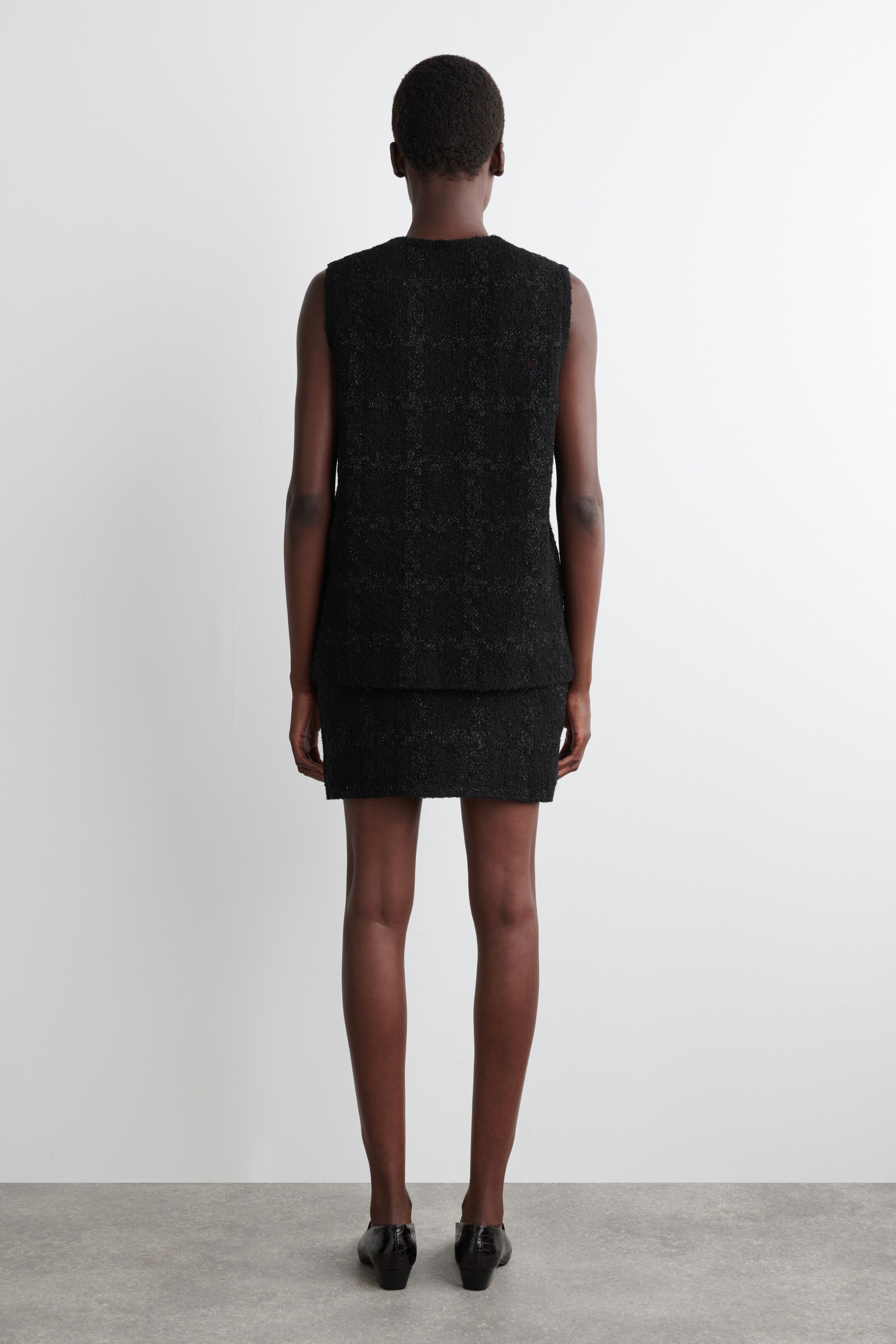 Grotere afbeelding bekijken: Een zwarte bouclé gebreide minirok, met een subtiel vensterruitpatroon en metallic draden, wordt van achteren gepresenteerd. Het is gestyled als een co-ord met een bijpassende mouwloze vest, waarbij beide stukken een reguliere pasvorm laten zien.