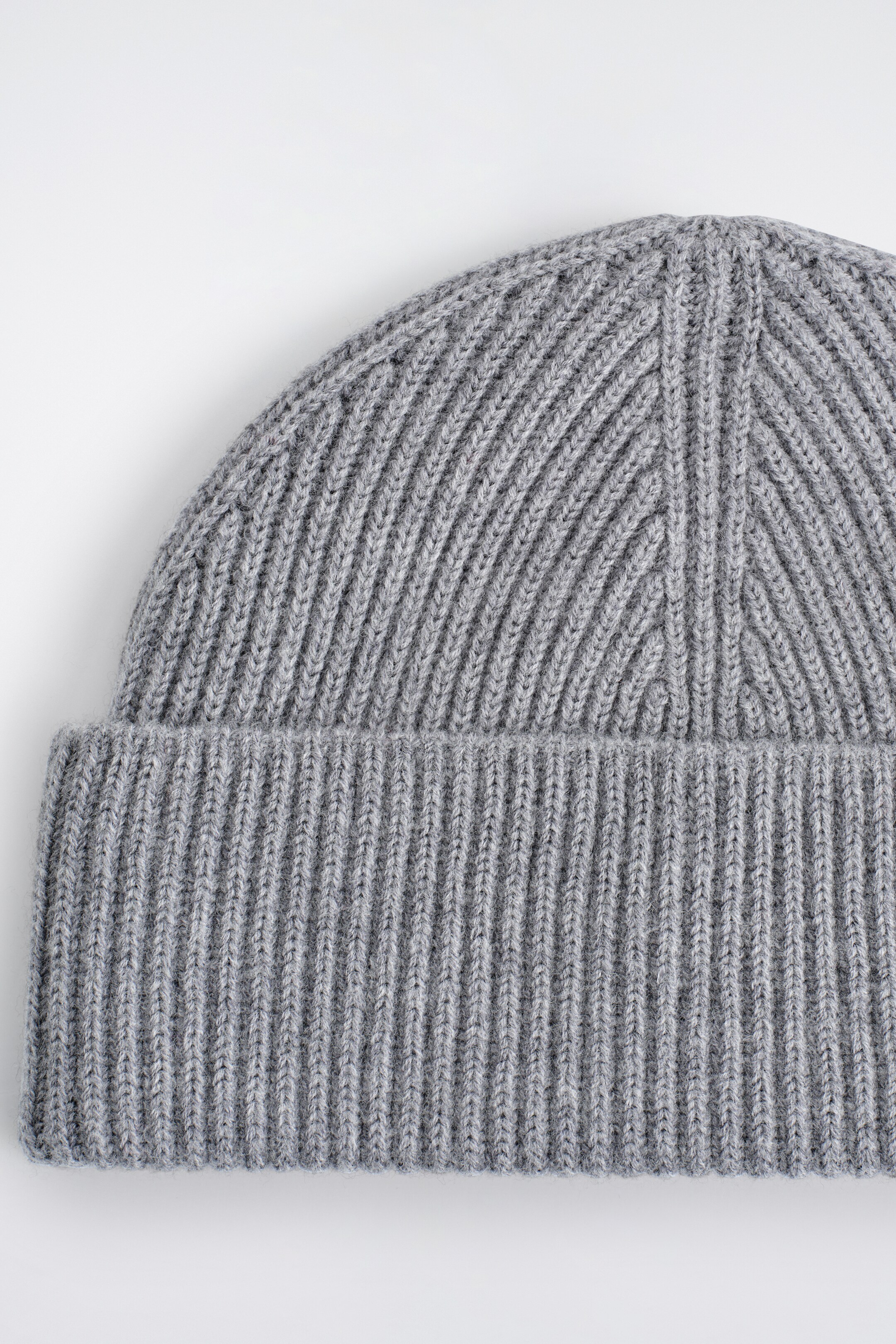 Ingrandisci l'immagine: Un berretto in maglia grigio presenta un'ampia piega a coste sul fondo. Anche la cima mostra una costruzione a coste, con il motivo a maglia che forma delle V verso l'alto.