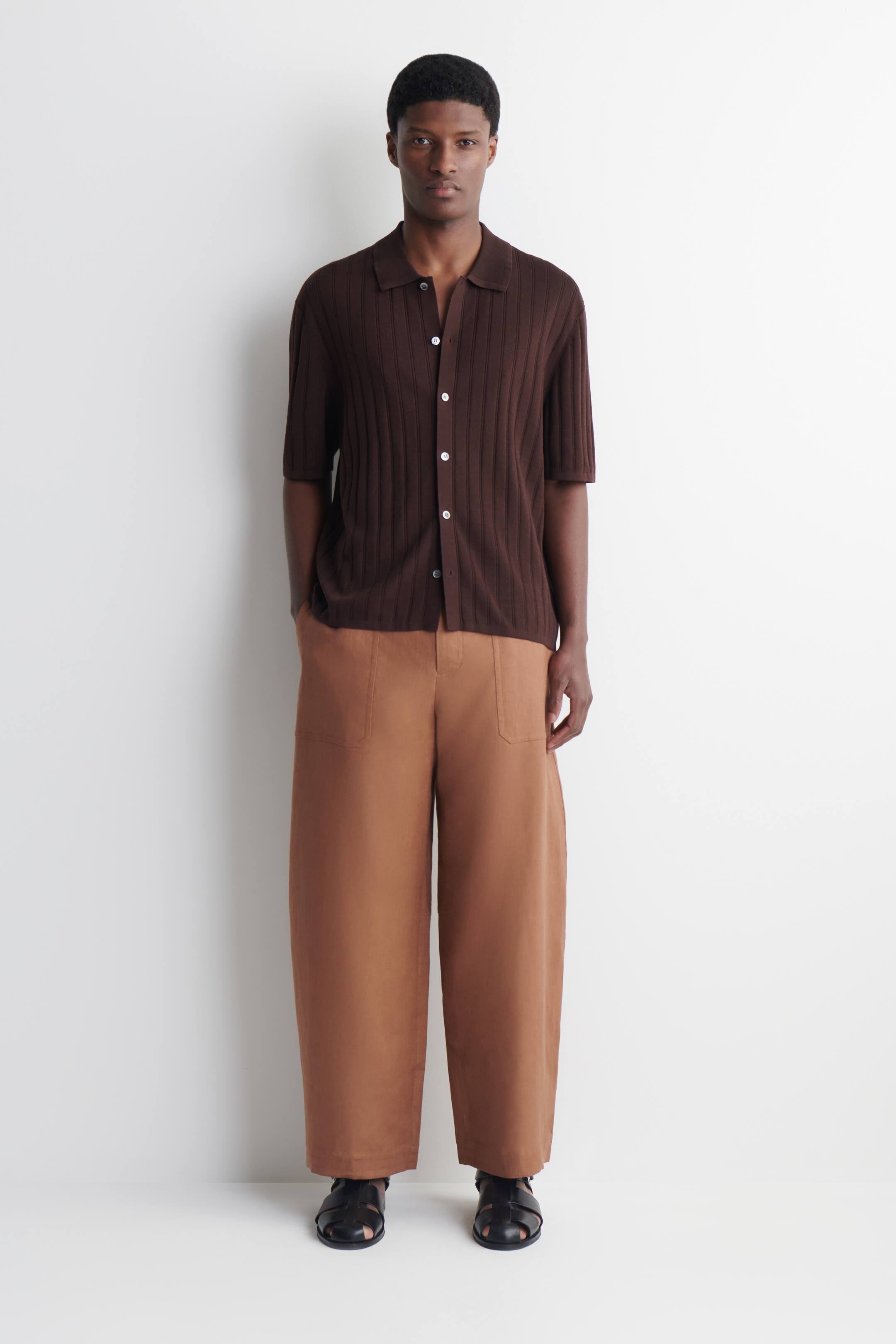 Grotere afbeelding bekijken: Een donkerbruine geribbelde gebreide poloshirt met korte mouwen toont een polokraag en een knoopsluiting aan de voorkant met witte knopen. Wijde beige broek met voorzakken en zwarte visserssandalen maken de outfit compleet, alles gepresenteerd met het gezicht naar voren.