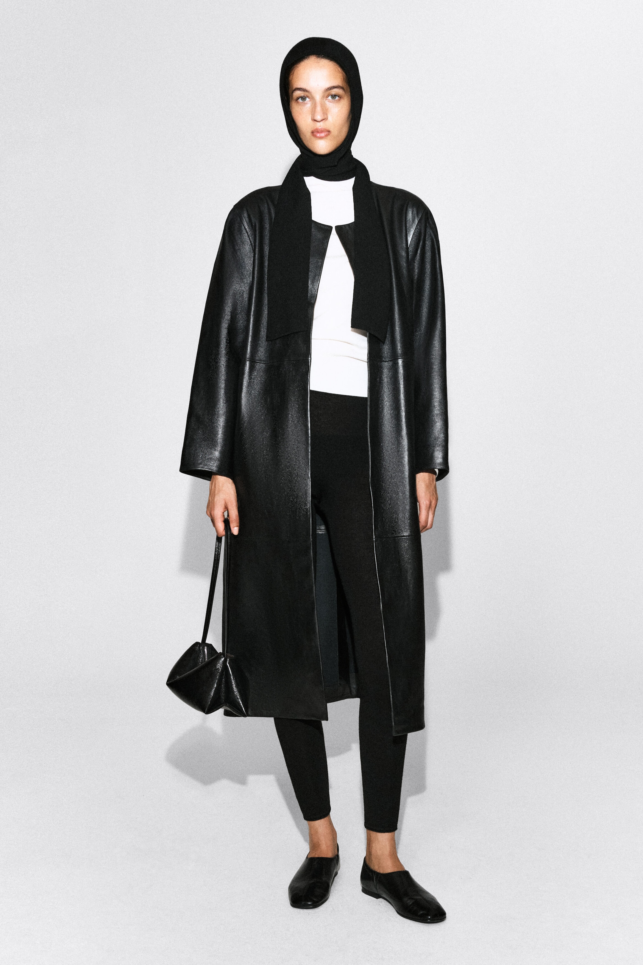 The model is wearing a black knitted hooded scarf that fits snugly over the head and drapes down the chest in two long panels. The accessory is styled over a white top and layered beneath a long, open black leather coat. The model is wearing a black knitted hooded scarf that fits snugly over the head and drapes down the chest in two long panels. The accessory is styled over a white top and layered beneath a long, open black leather coat.