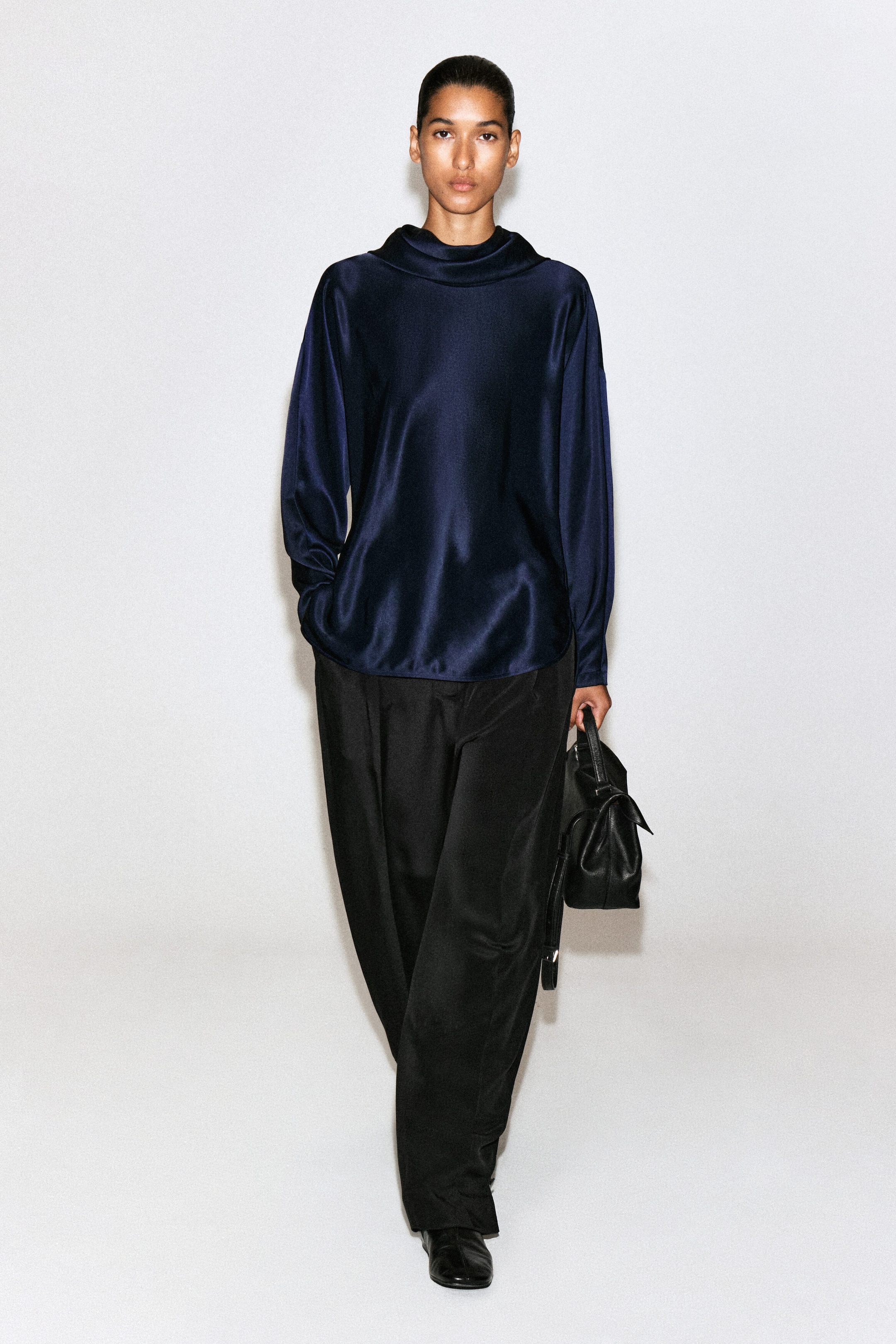 A cowl-neck satin blouse in navy blue is shown from the front, featuring a high draped neckline, long sleeves, and a relaxed silhouette. The blouse is hanging untucked over wide-leg black trousers, showcasing its fluid drape and lustrous fabric.