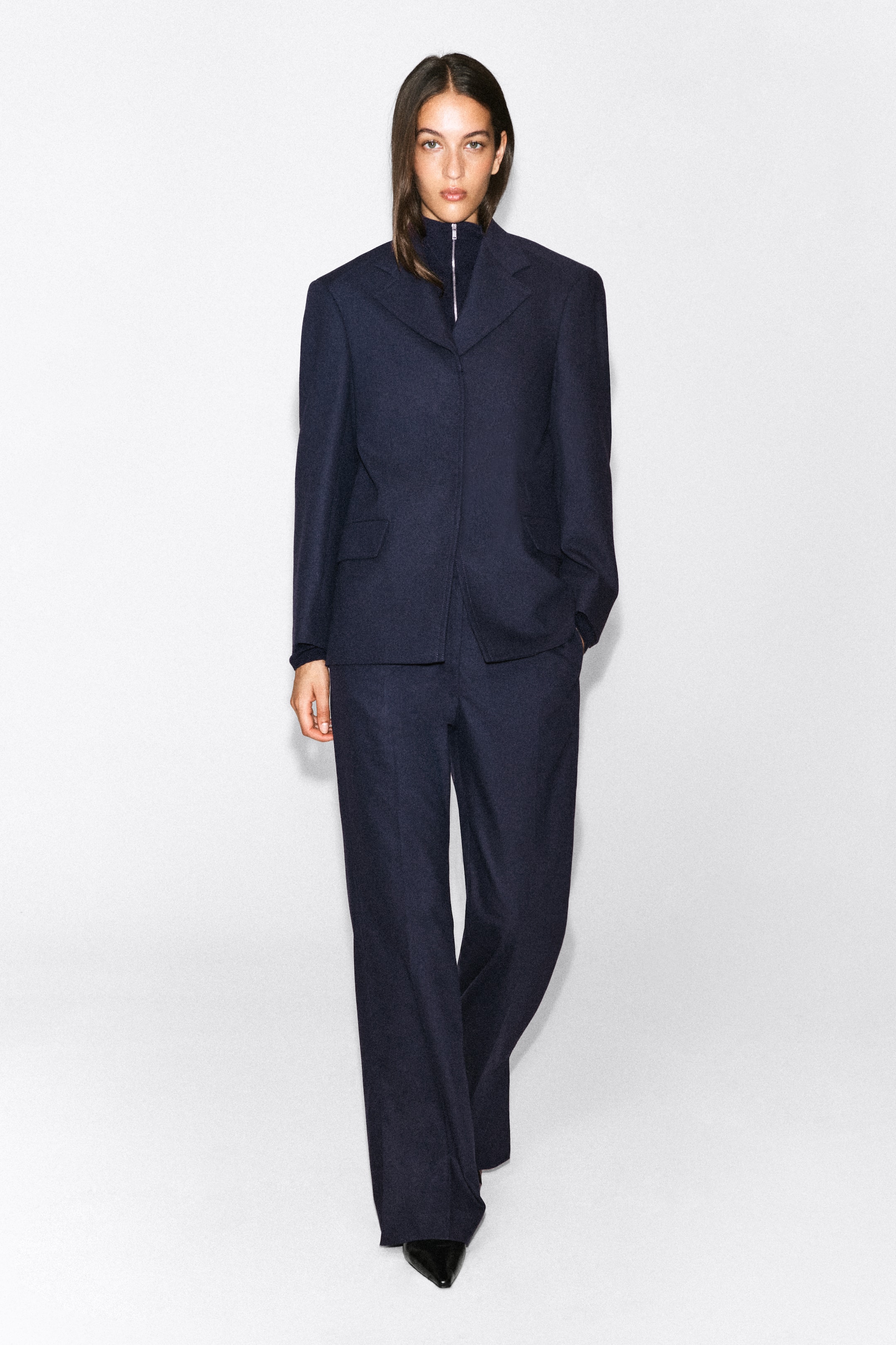 Agrandir l'image: Un pantalon évasé en mélange de laine bleu marine foncé est porté par le mannequin, avec une silhouette large et des plis marqués sur le devant. Le pantalon est associé à un blazer structuré assorti et à un haut sombre zippé à col montant.