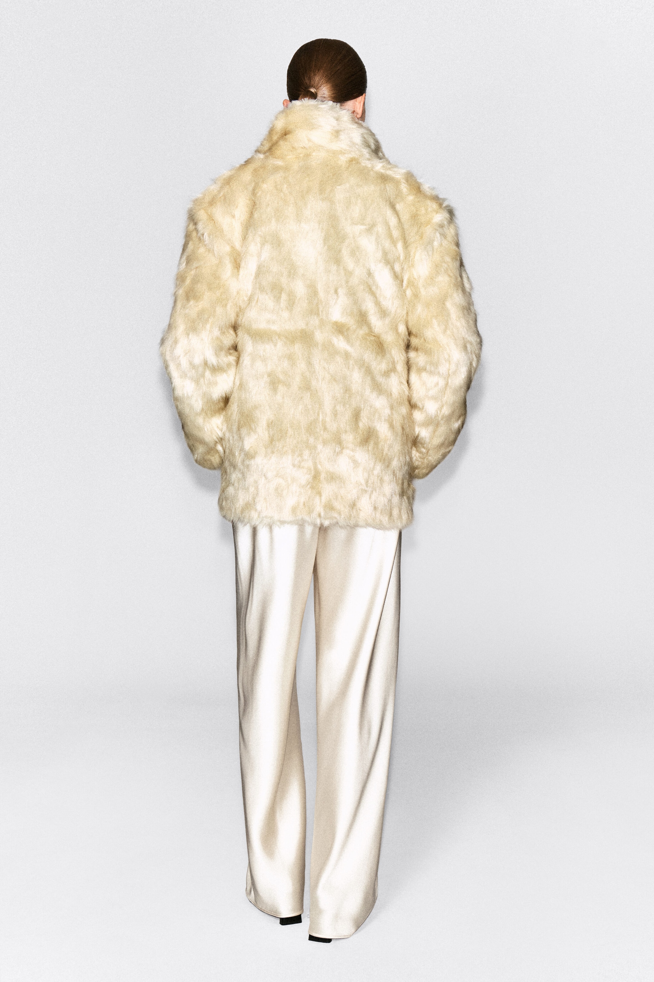 A cream-colored faux-fur jacket is being worn by the model, who is standing with their back to the camera. This relaxed-fit jacket features a plush texture and a high collar, and is styled with cream-colored, wide-leg satin trousers.