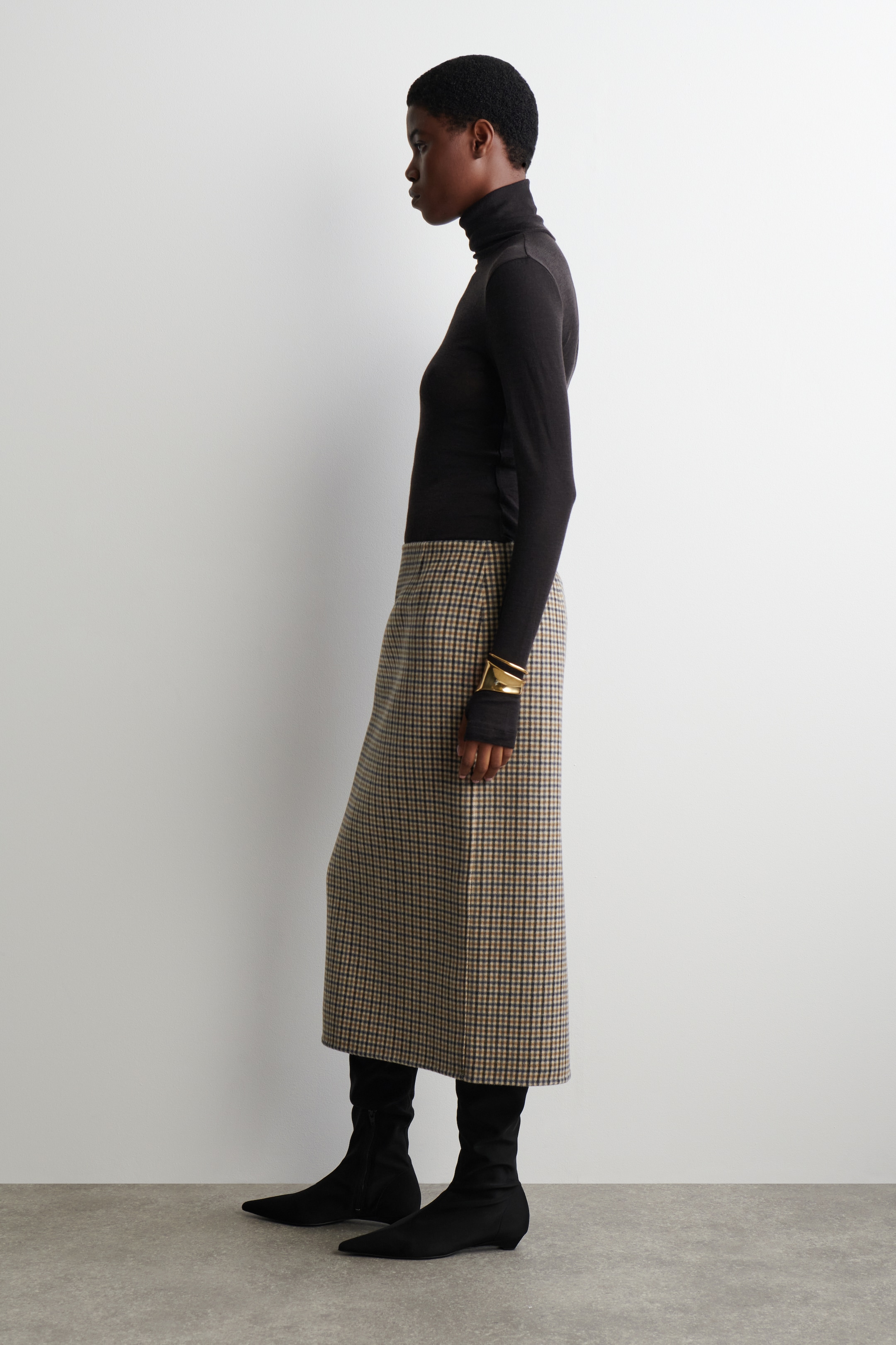 Agrandir l'image: Un col roulé noir ajusté, une jupe midi à carreaux marron et beige, et des bottines noires à bout pointu sont présentés sur un mannequin de profil tourné vers la droite. Le col roulé présente un col haut, et la jupe affiche un motif à petits carreaux, avec un large bracelet manchette doré accessoirisant le poignet droit.