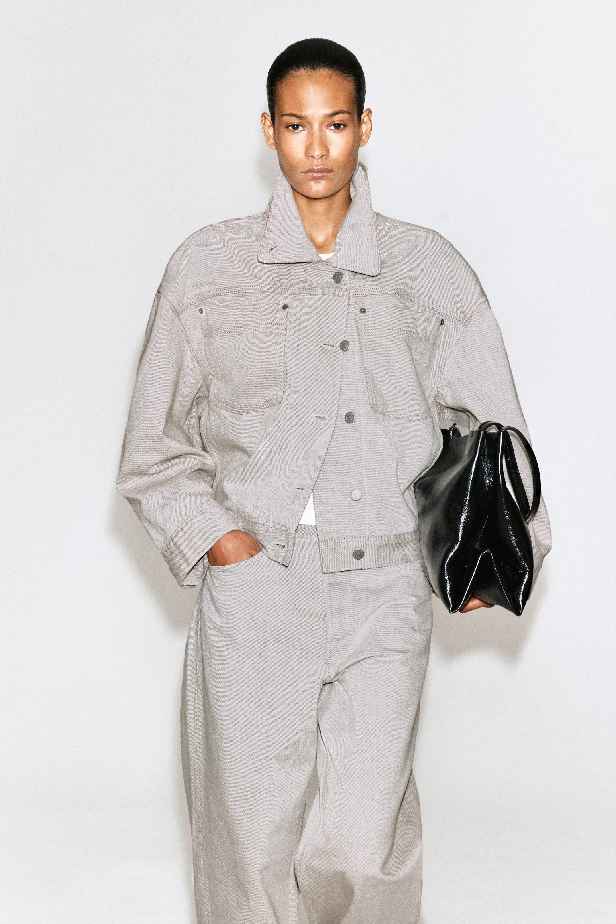 View larger image: A light grey denim jacket, featuring a stand-up collar, button-front closure, chest flap pockets and visible stitching, is styled with matching wide-leg trousers. A hand rests in a trouser pocket while a shiny black sculptural handbag, with a distinctive folded design, is held.