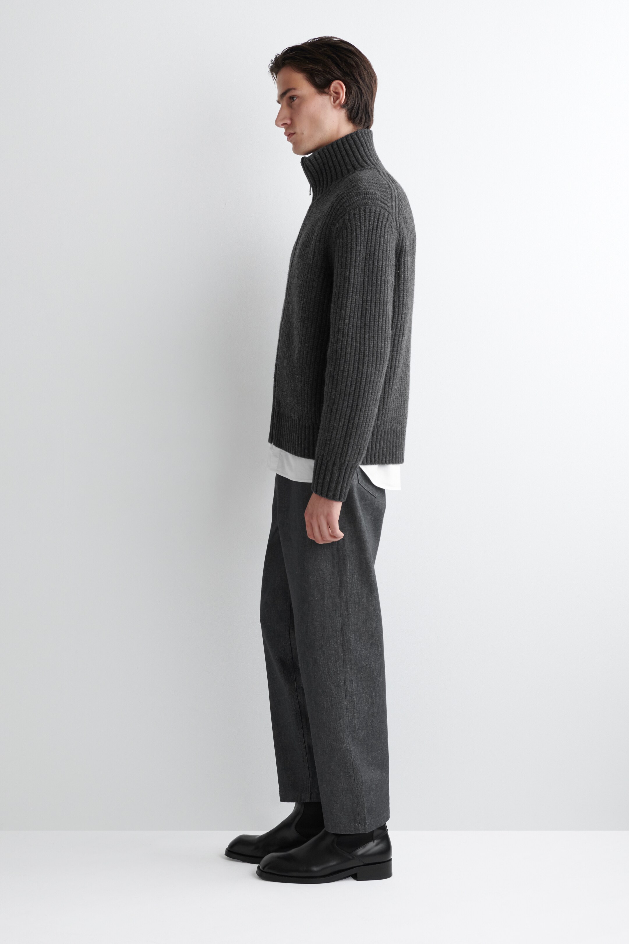 Agrandir l'image: Un pull à col quart-zip gris foncé est présenté de profil, avec une texture tricotée côtelée et un col montant avec une fermeture éclair visible. Un t-shirt blanc est superposé en dessous, visible à l'ourlet.