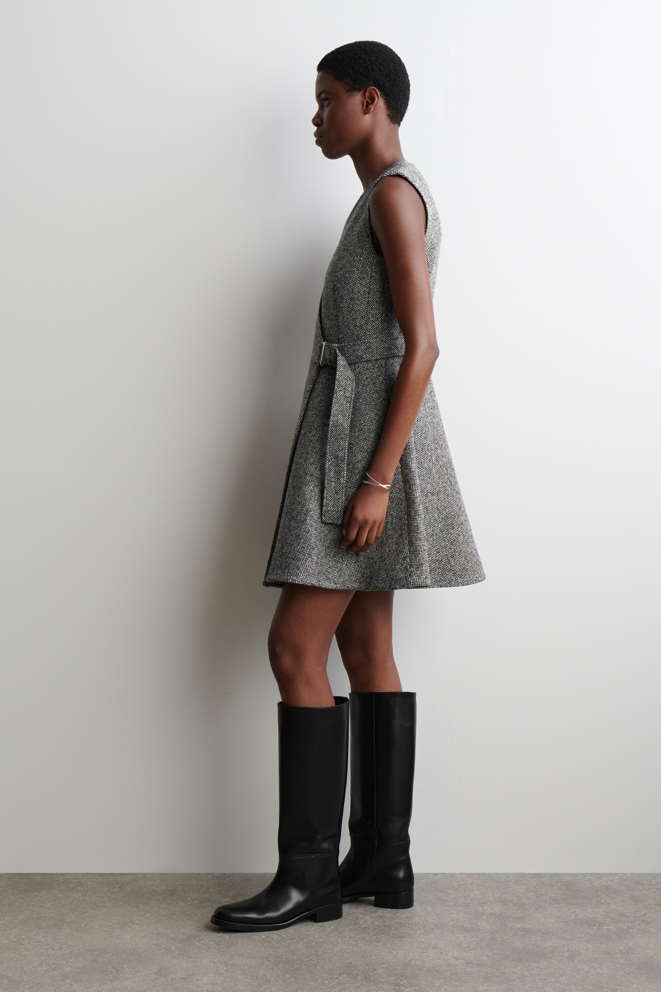 Se større billede: En ærmeløs, grå tweed wrap-kjole præsenteres fra en sideprofil, den har en bælte i taljen med et rektangulært spænde og en udsvingt nederdel, der slutter over knæet. Den bliver stylet med sorte knælange støvler.