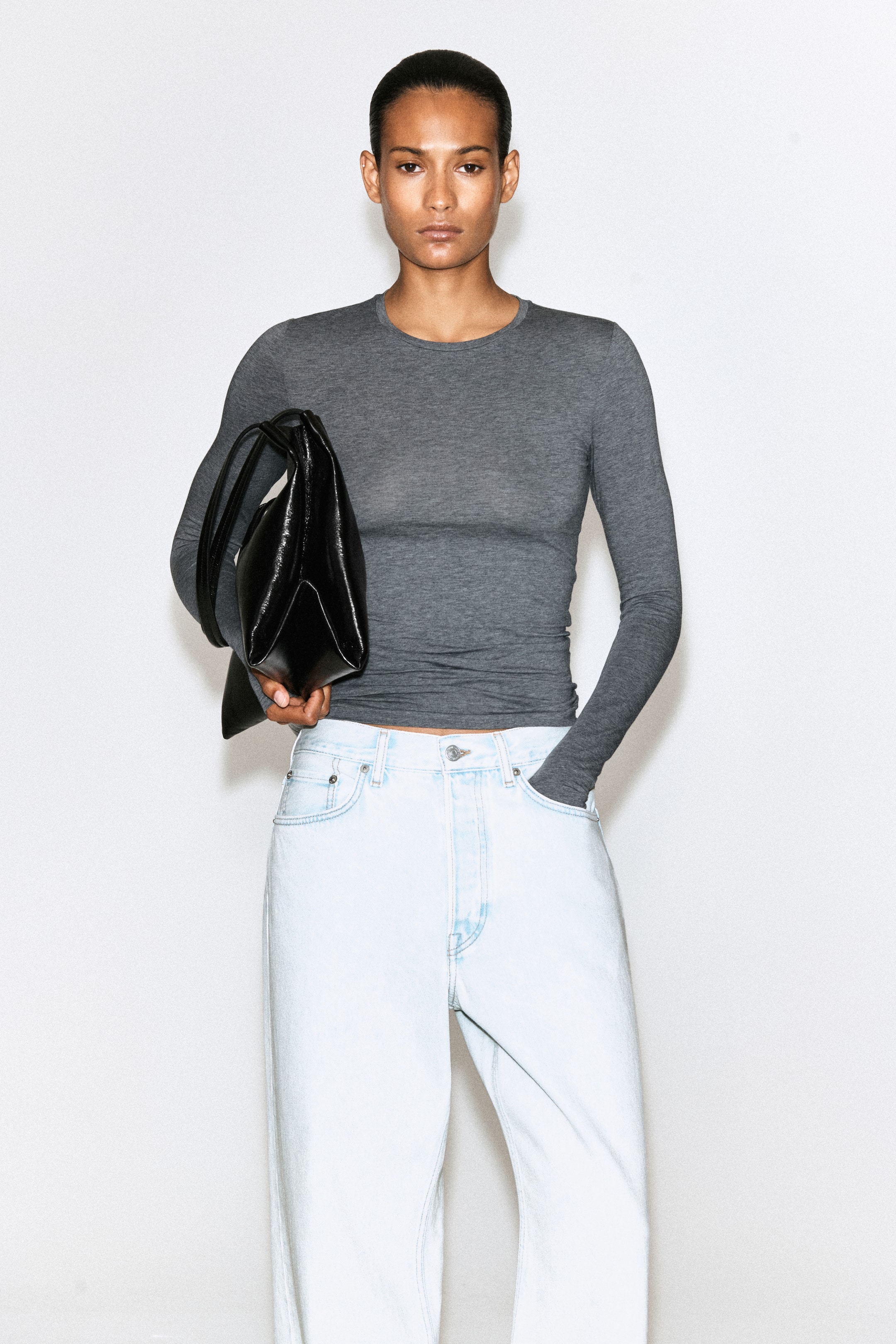 View larger image: The model is wearing a dark grey, slim-fit, long-sleeved crew-neck top. The model’s right thumb is through a thumbhole cuff, and the left sleeve covers their hand as it rests in a jean pocket.