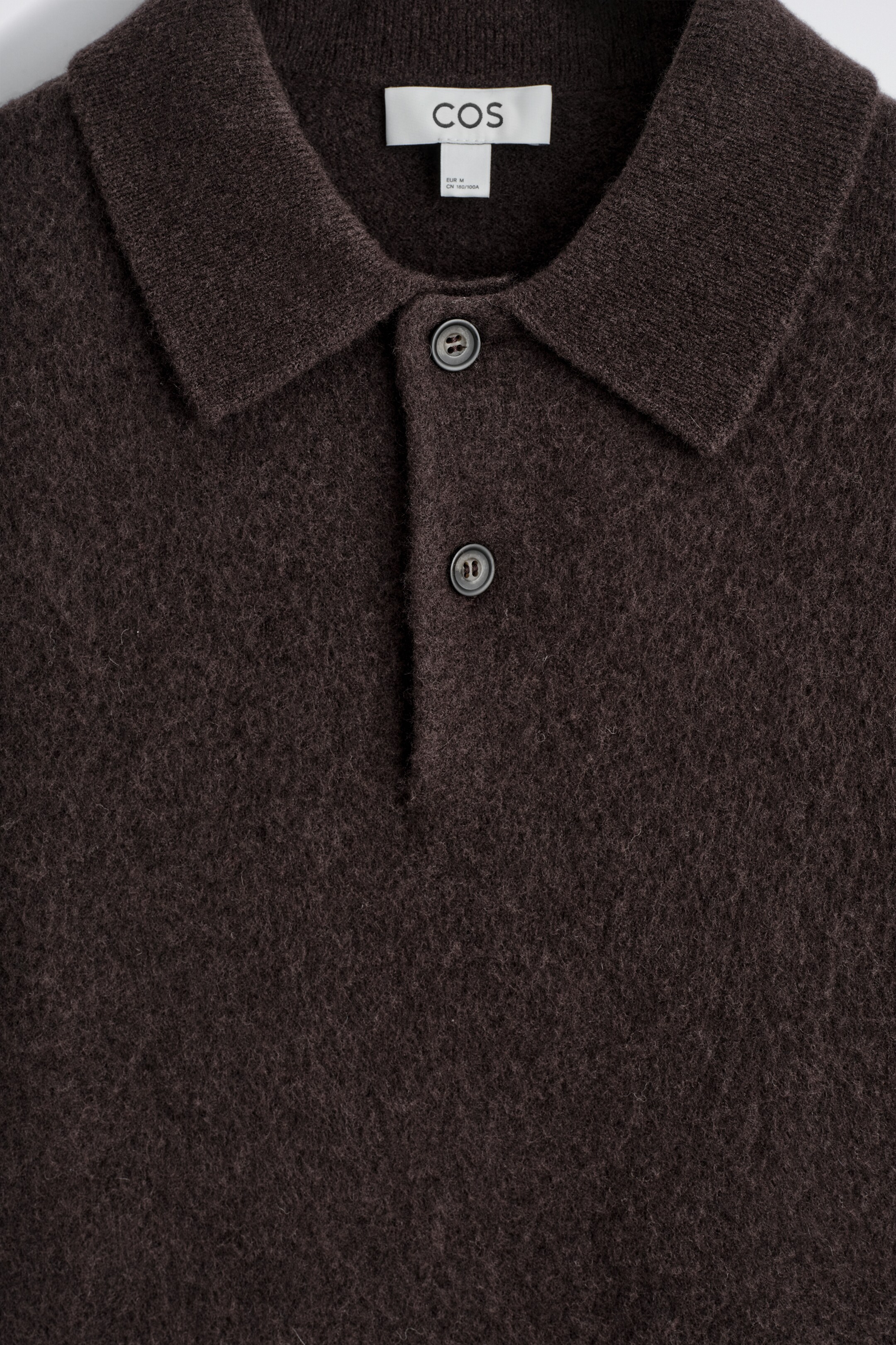 Agrandir l'image: Un polo marron foncé met en valeur sa texture visiblement brossée à l'avant, avec un col classique et une patte de boutonnage à deux boutons ronds et foncés.