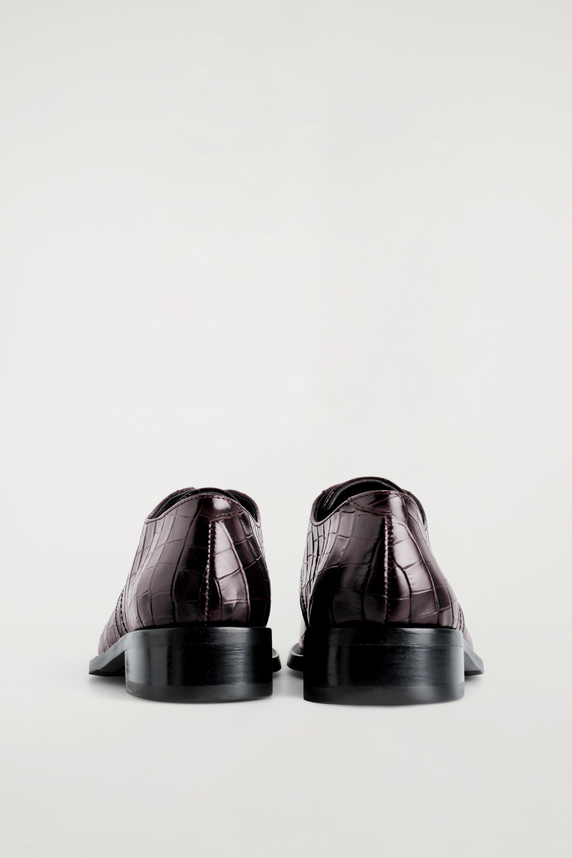 Visualizza immagine più grande: Due paia di scarpe Derby color espresso di COS vengono presentate dal retro, mettendo in mostra la loro texture in pelle con stampa coccodrillo. Presentano tacchi bassi e scuri a blocco.