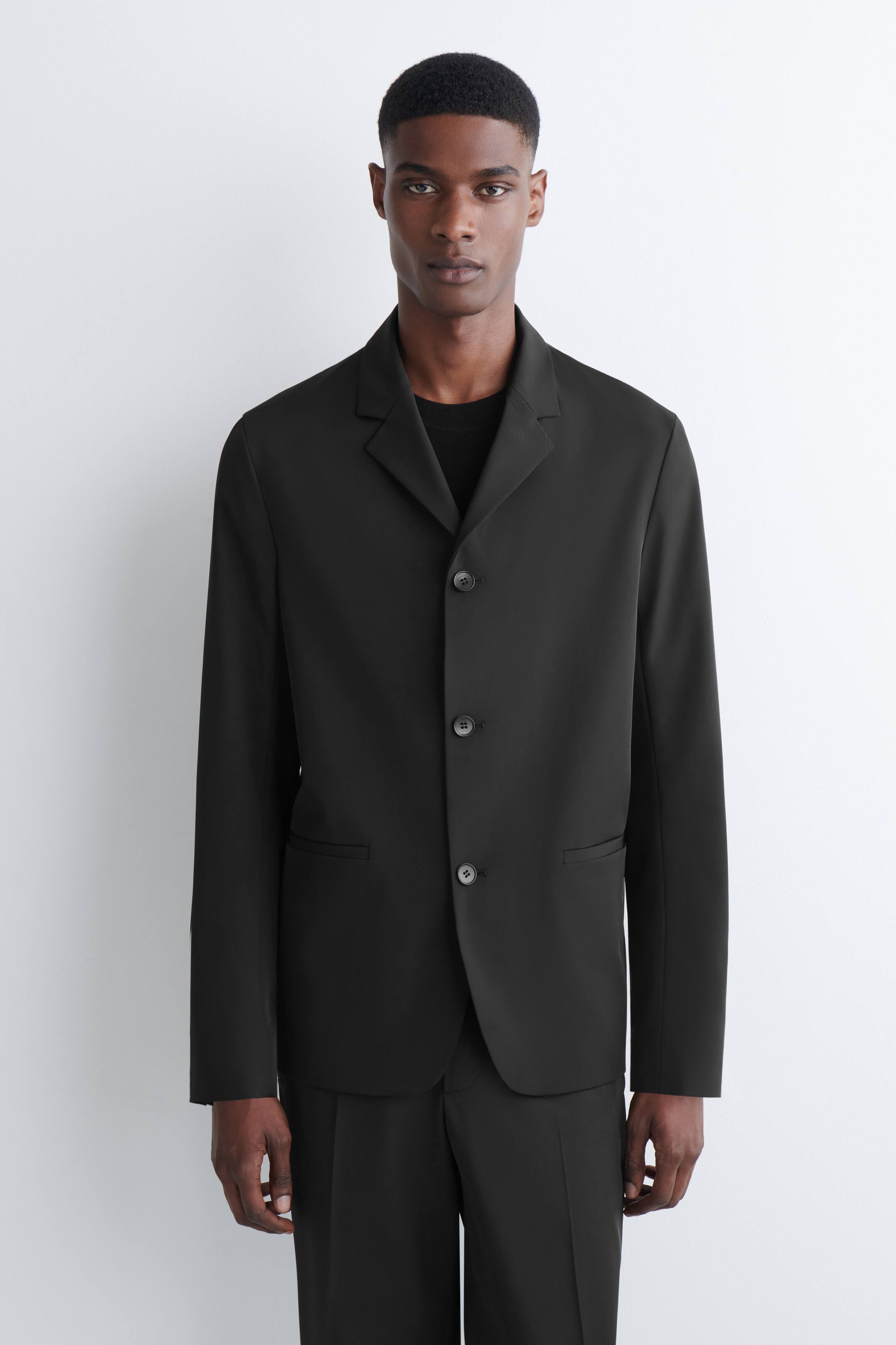 Grösseres Bild ansehen: Ein schwarzer Blazer wird von vorne präsentiert, mit Kerbrevers, einem Drei-Knopf-Verschluss, einer einzelnen Leistentasche auf der Brust und zwei Pattentaschen an den Hüften. Er wird über einem schwarzen T-Shirt und passenden schwarzen Hosen gestylt.