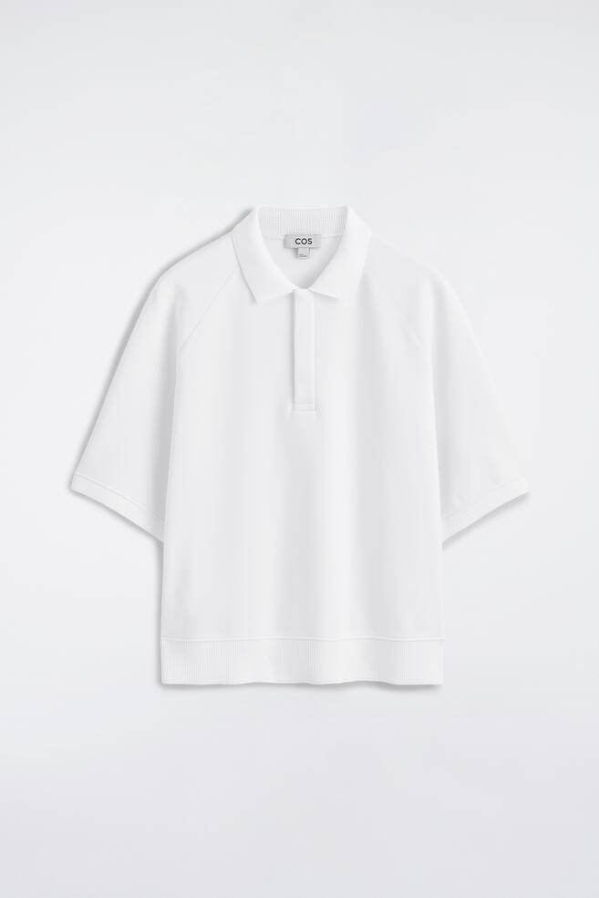 Das weiße Poloshirt ist von vorne präsentiert und verfügt über einen gerippten Kragen, eine kurze Knopfleiste in der Mitte sowie kurze Raglanärmel. Gerippte Bündchen schließen die Ärmel ab, und ein breiter gerippter Saum rundet den unteren Abschluss ab. - 1