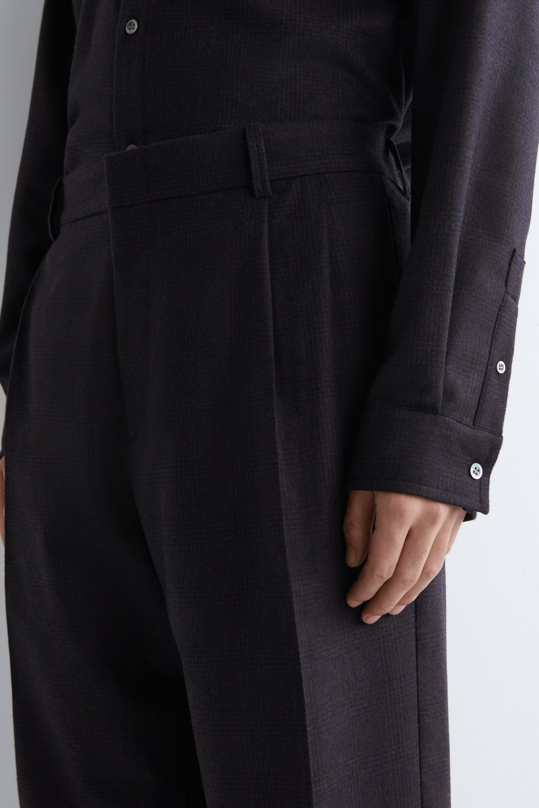 Agrandir l'image: Un pantalon foncé affiche un motif de micro-carreaux subtil, avec des plis sur le devant et une ceinture structurée, présenté de face et en biais. Une chemise à manches longues assortie est soigneusement rentrée, ses poignets, chacun fermé par deux boutons de couleur claire, sont visibles au poignet où une main repose.