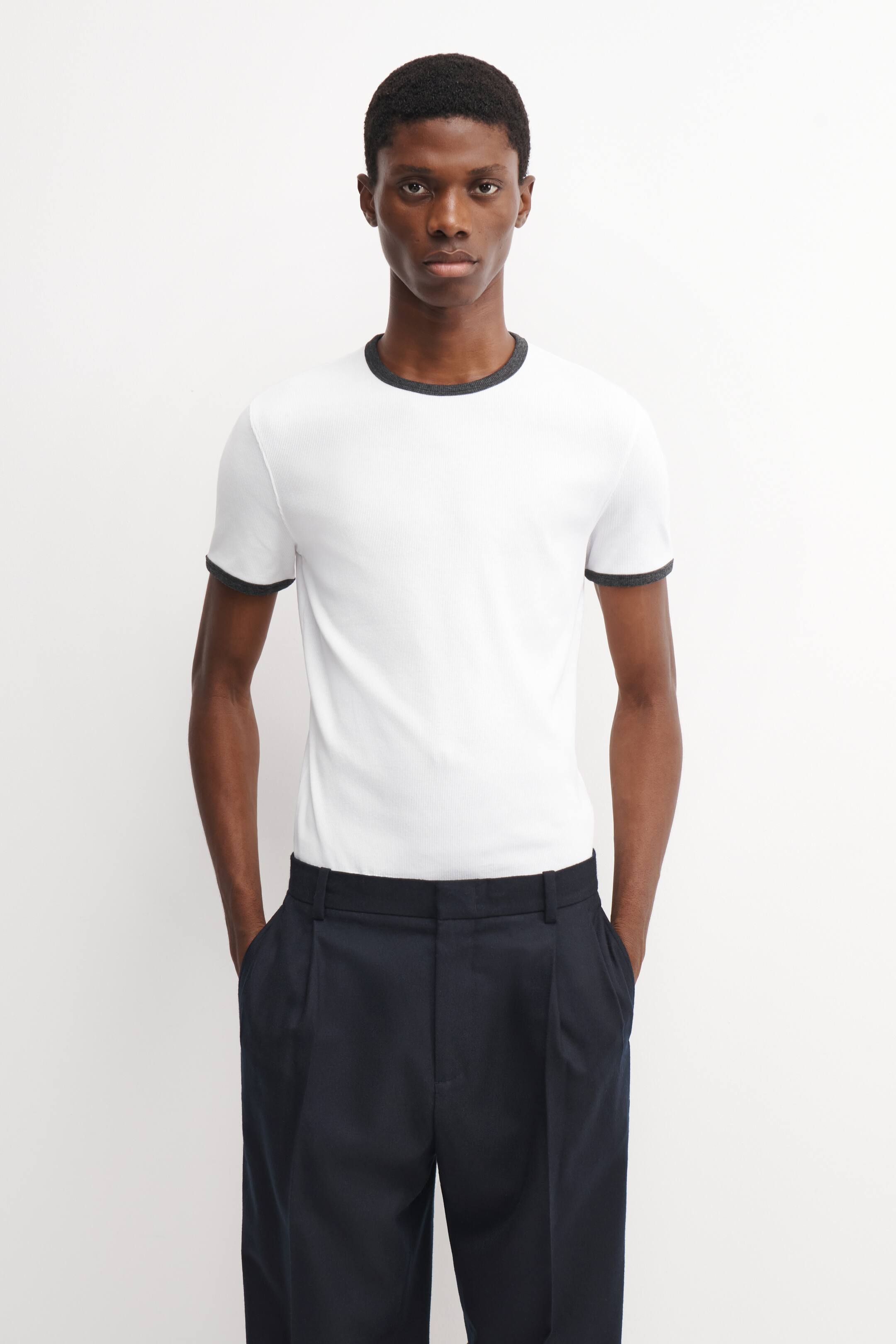 Ver imagen más grande: Una camiseta de algodón acanalado y entallada en blanco, con cuello redondo en contraste de color gris oscuro y puños de las mangas a juego, la lleva puesta la modelo. La camiseta está por dentro de un pantalón oscuro plisado.