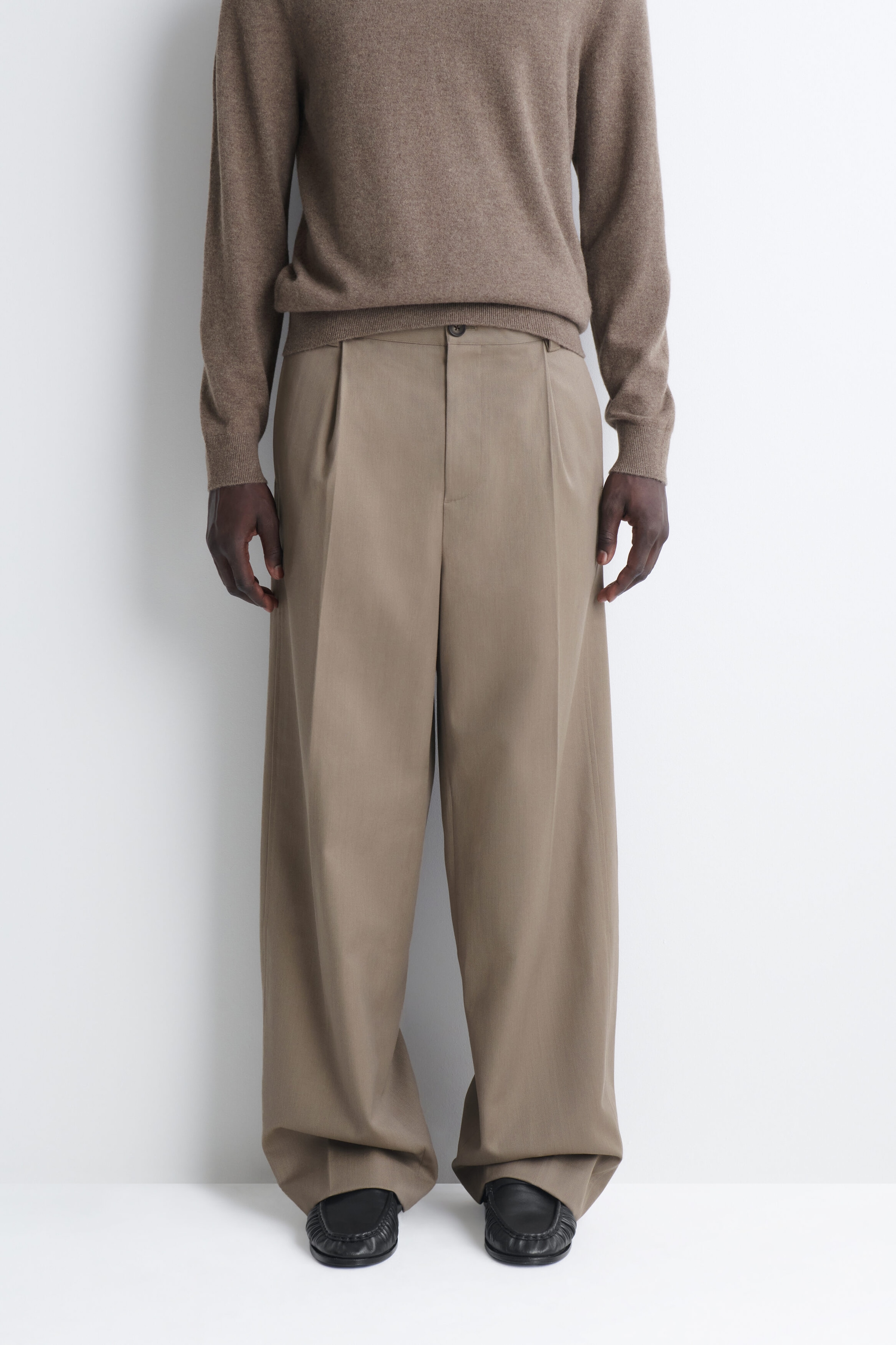 Unos pantalones beige de pernera ancha presentan una silueta relajada y fluida con un único pliegue delantero y un marcado pliegue central a lo largo de cada pierna. Los pantalones se asientan a la cintura, por debajo de un jersey de punto marrón claro, y caen ligeramente sobre unos zapatos negros. Unos pantalones beige de pernera ancha presentan una silueta relajada y fluida con un único pliegue delantero y un marcado pliegue central a lo largo de cada pierna. Los pantalones se asientan a la cintura, por debajo de un jersey de punto marrón claro, y caen ligeramente sobre unos zapatos negros.