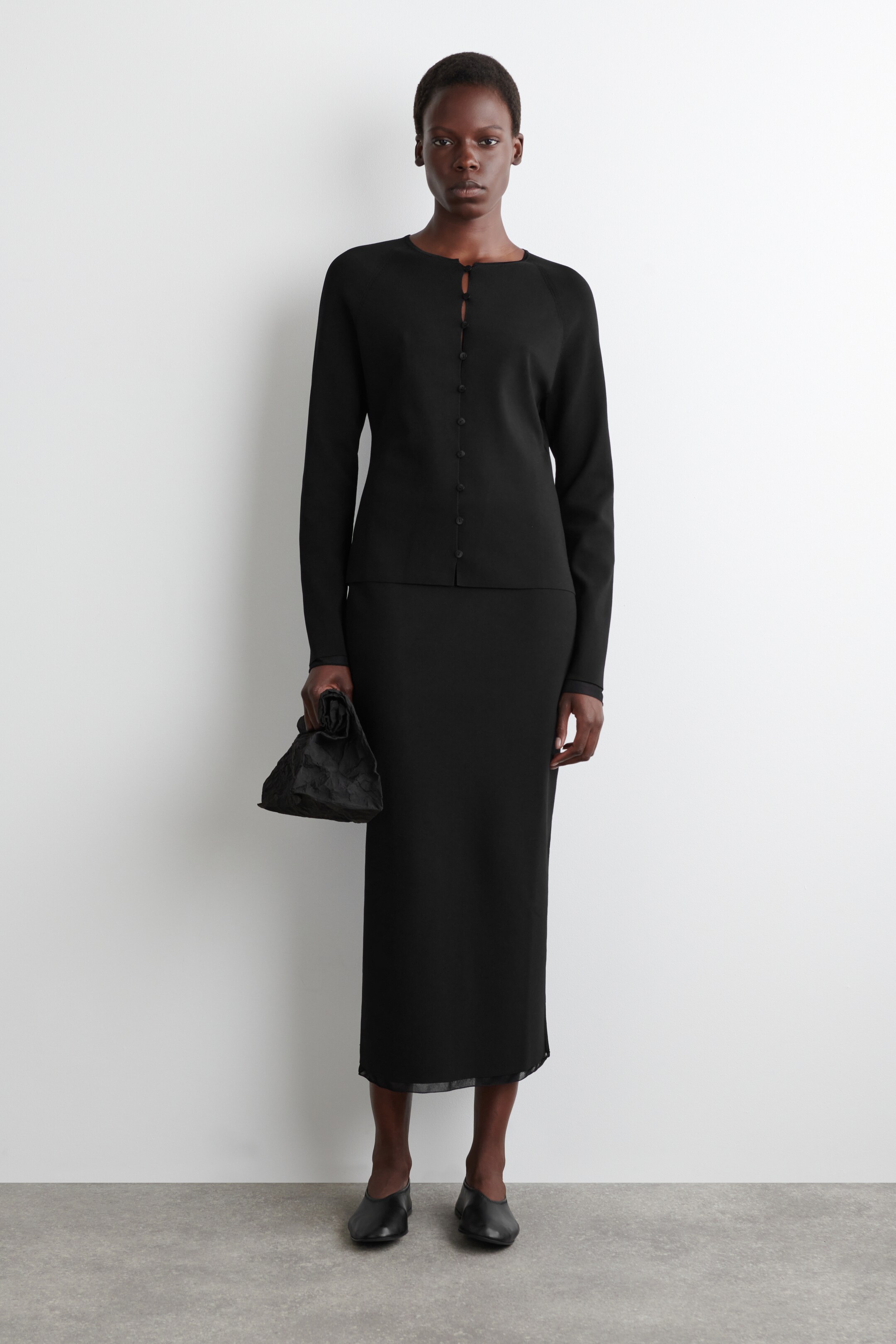 View larger image: A black long-sleeved cardigan with a crew neck and full button-front closure is being worn over a coordinating black midi skirt featuring a subtle sheer trim at the hem, all facing forward. A small black ruched handbag is being held, and black flat shoes are being worn.