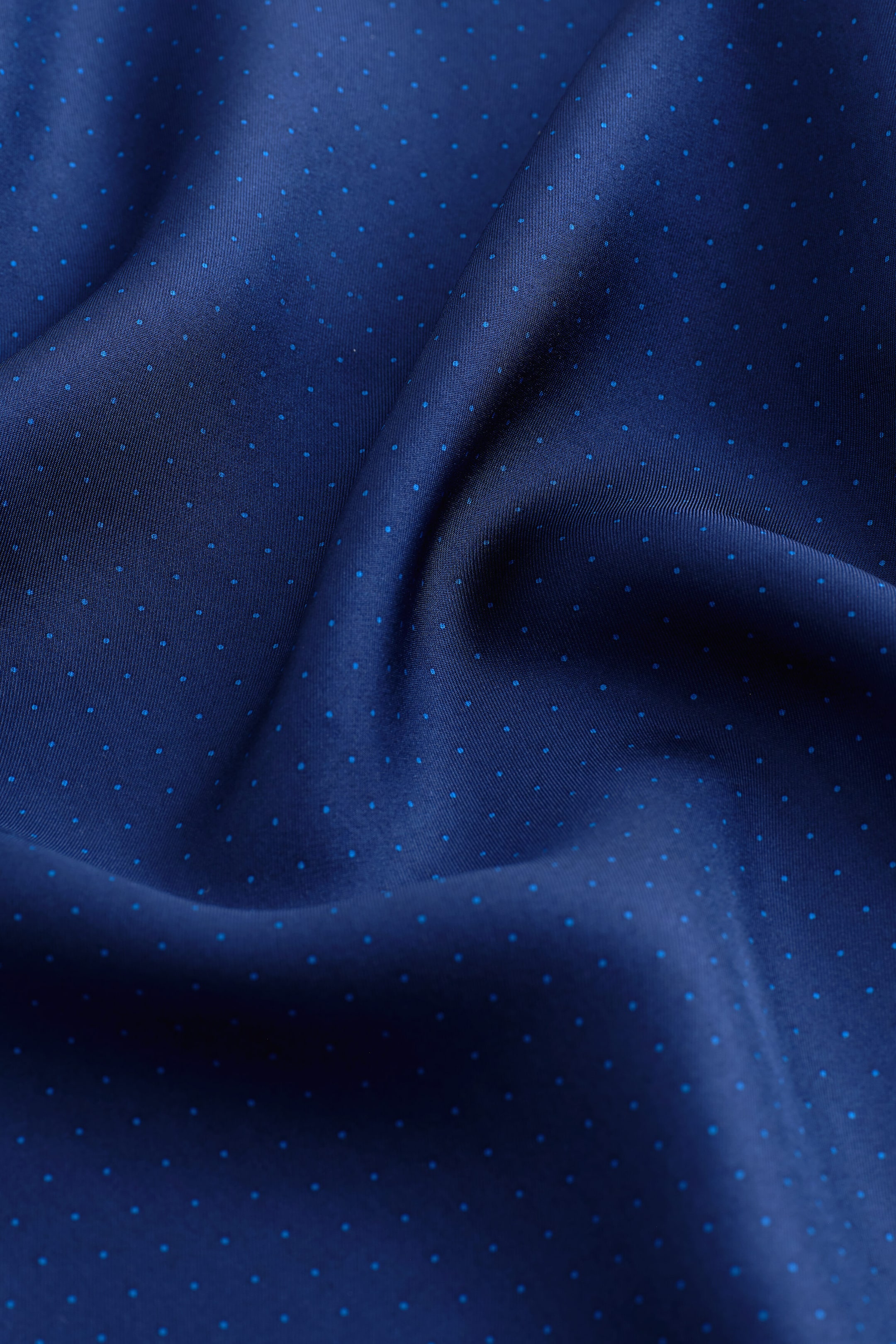 A polka-dot silk pocket square displays a navy blue base adorned with a pattern of tiny light blue pin dots. The smooth, lustrous fabric drapes in soft, rippling folds to showcase its texture and subtle sheen.