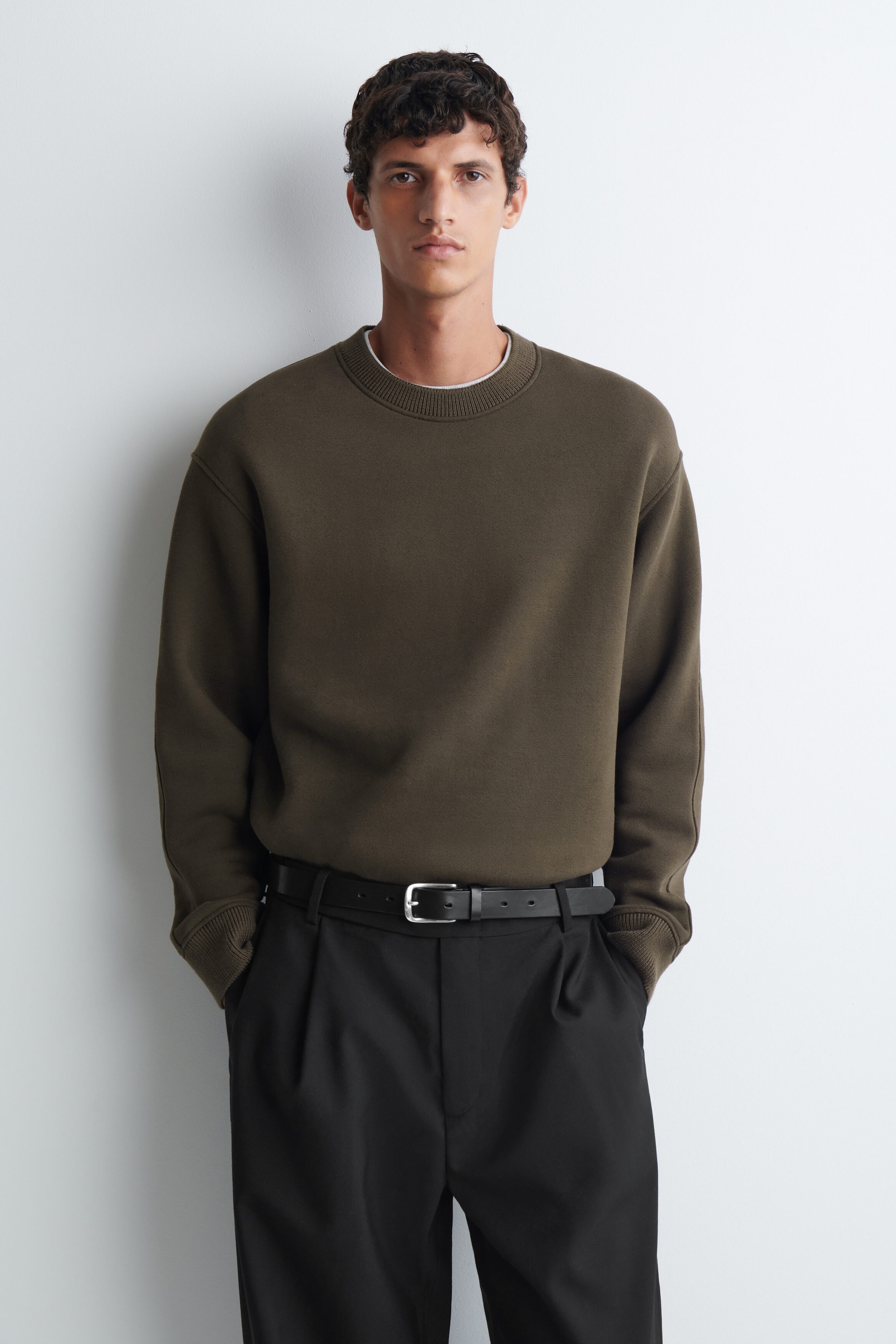 Le mannequin porte un sweatshirt en jersey de coton et de laine vert olive, doté d’un col rond côtelé et de poignets côtelés. Ce sweatshirt à coupe décontractée est rentré dans un pantalon plissé noir maintenu par une ceinture en cuir noir. Le mannequin porte un sweatshirt en jersey de coton et de laine vert olive, doté d’un col rond côtelé et de poignets côtelés. Ce sweatshirt à coupe décontractée est rentré dans un pantalon plissé noir maintenu par une ceinture en cuir noir.
