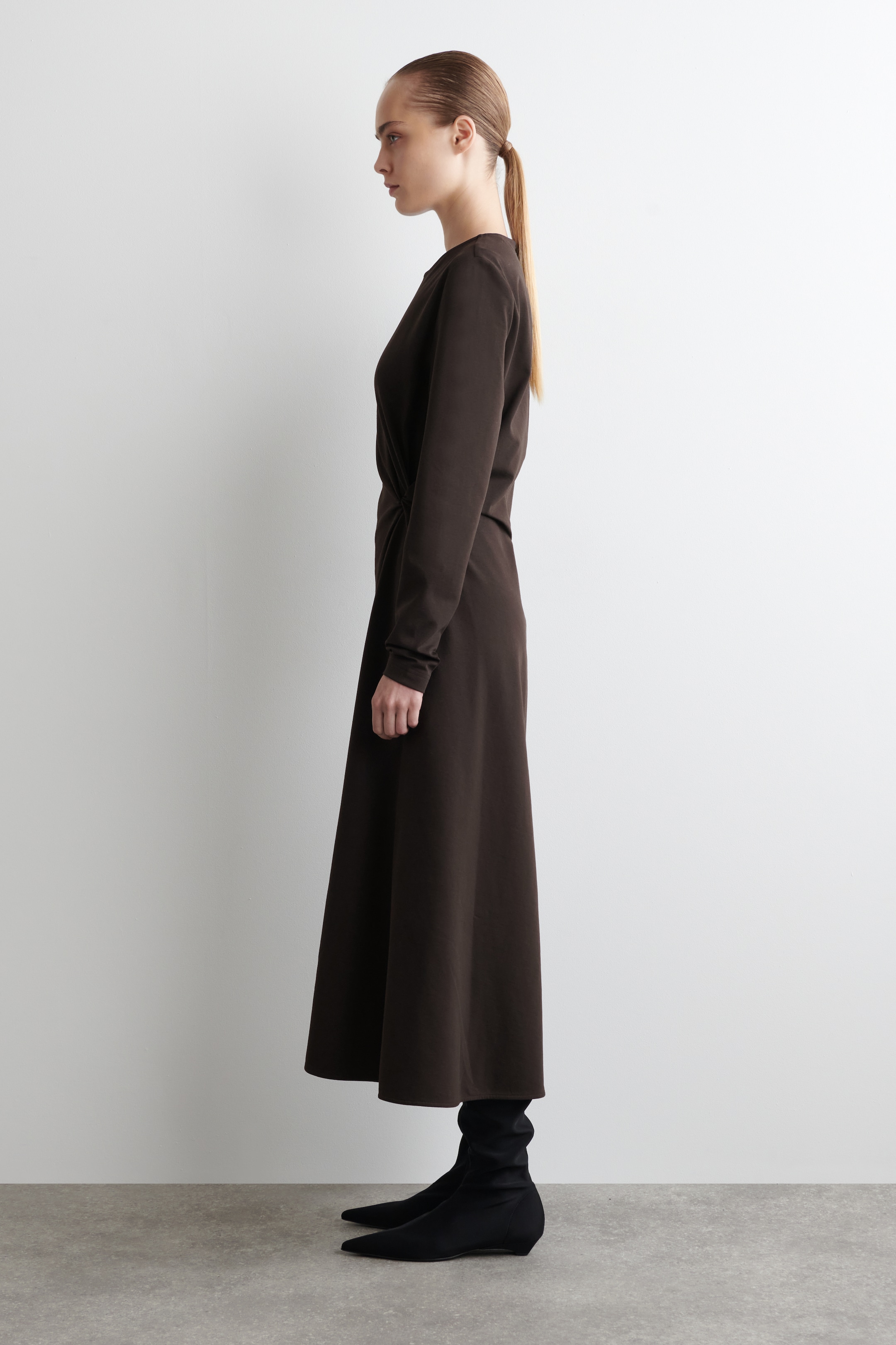 Agrandir l'image: Une robe marron foncé à manches longues est présentée de profil, vers la droite. Elle est dotée d'un col rond, d'un détail froncé à la taille sur le côté, et d'une jupe évasée qui tombe au milieu du mollet par-dessus des bottes noires.