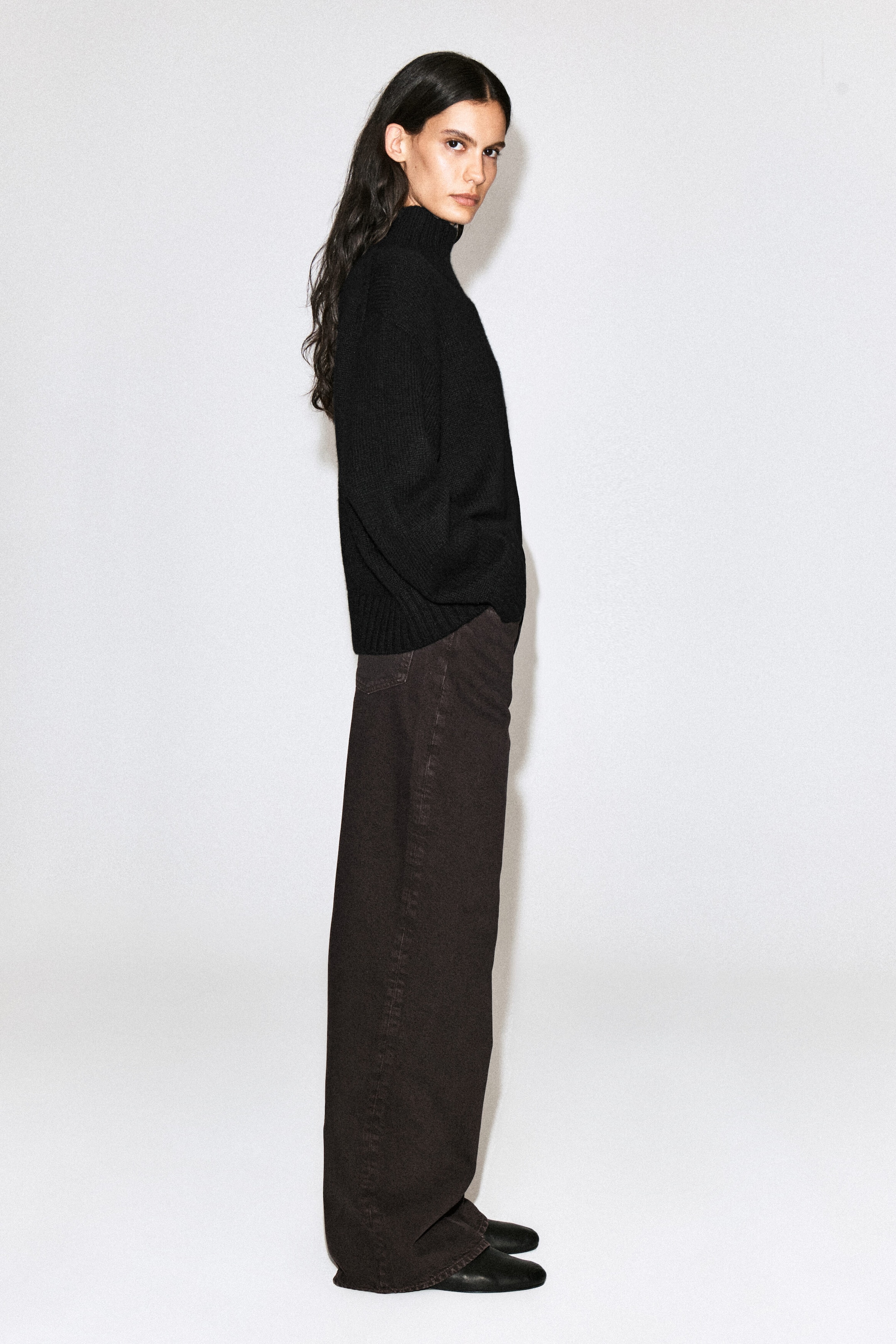The model is wearing dark brown denim wide-leg jeans while standing in profile. The jeans feature a relaxed, wide-leg silhouette and are styled with a black turtleneck sweater and black shoes. The model is wearing dark brown denim wide-leg jeans while standing in profile. The jeans feature a relaxed, wide-leg silhouette and are styled with a black turtleneck sweater and black shoes.