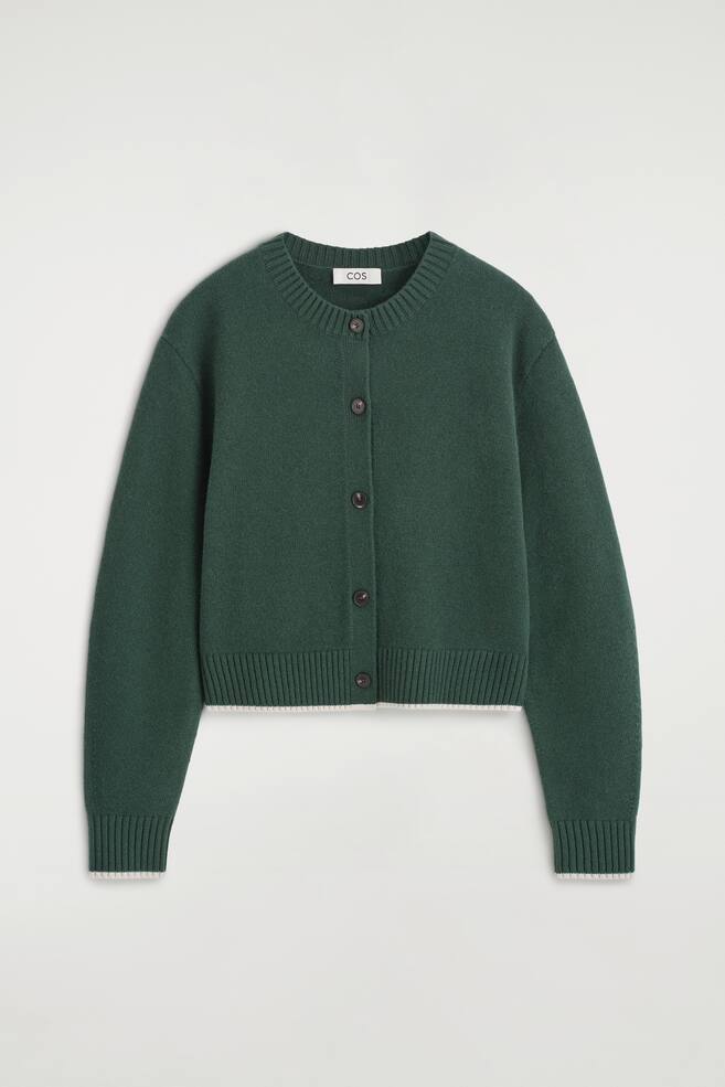 En mÃ¸rkegrÃ¸n cardigan i merinould er vendt frontalt og fremhÃ¦ver en knaplukning foran med fem mÃ¸rke knapper. Den har rund hals med ribkant, ribbede manchetter og ribkant forneden, alle afsluttet med en tynd, kontrasterende hvid kant. - 1