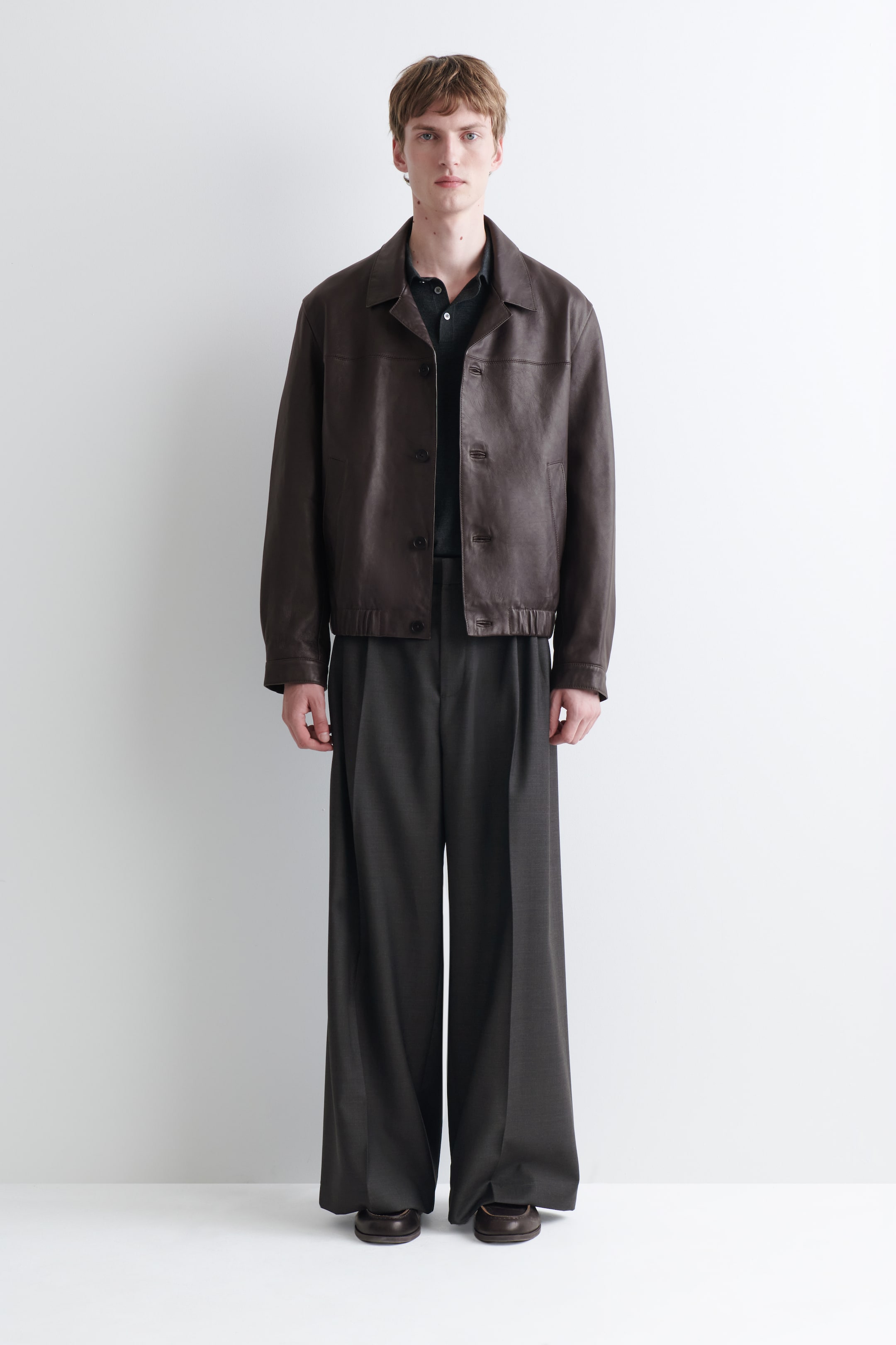Agrandir l'image: Une veste en cuir marron avec col, dotée de boutons sur le devant et d’un ourlet élastiqué, est portée par le mannequin. Elle est superposée à un polo foncé.