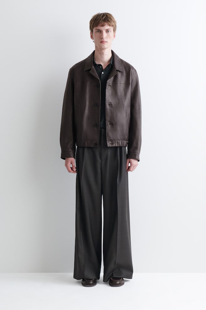 Une veste en cuir marron avec col, dotée de boutons sur le devant et dâun ourlet élastiqué, est portée par le mannequin. Elle est superposée à un polo foncé. - 1