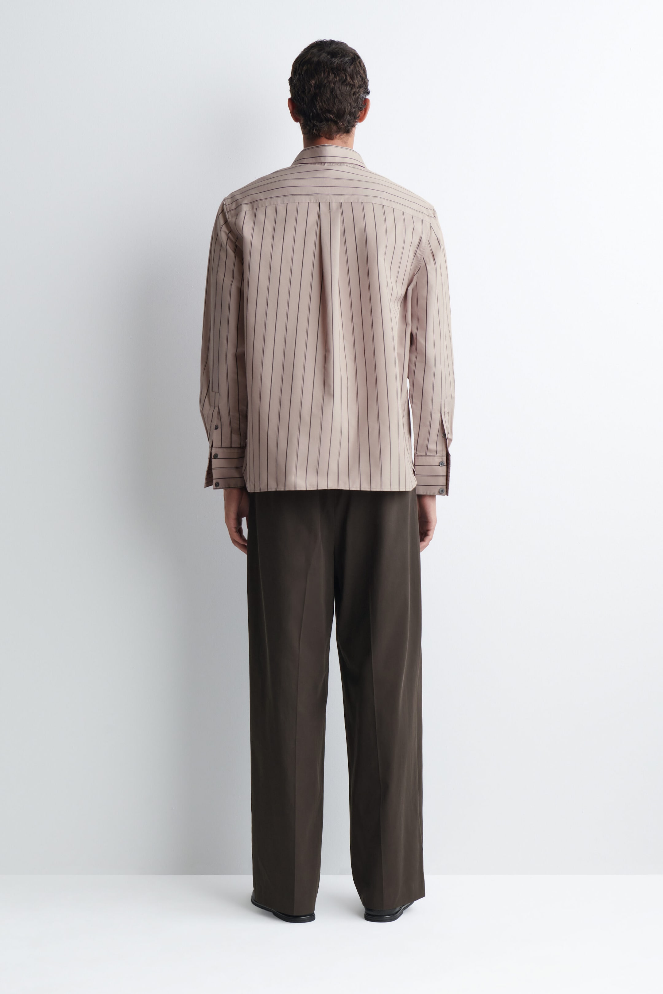 A striped structured cotton-silk shirt in beige with thin, dark vertical stripes is being worn by the model, shown from a back-facing view. The long-sleeved shirt features a back yoke with a central box pleat and is styled with dark brown wide-leg trousers. A striped structured cotton-silk shirt in beige with thin, dark vertical stripes is being worn by the model, shown from a back-facing view. The long-sleeved shirt features a back yoke with a central box pleat and is styled with dark brown wide-leg trousers.