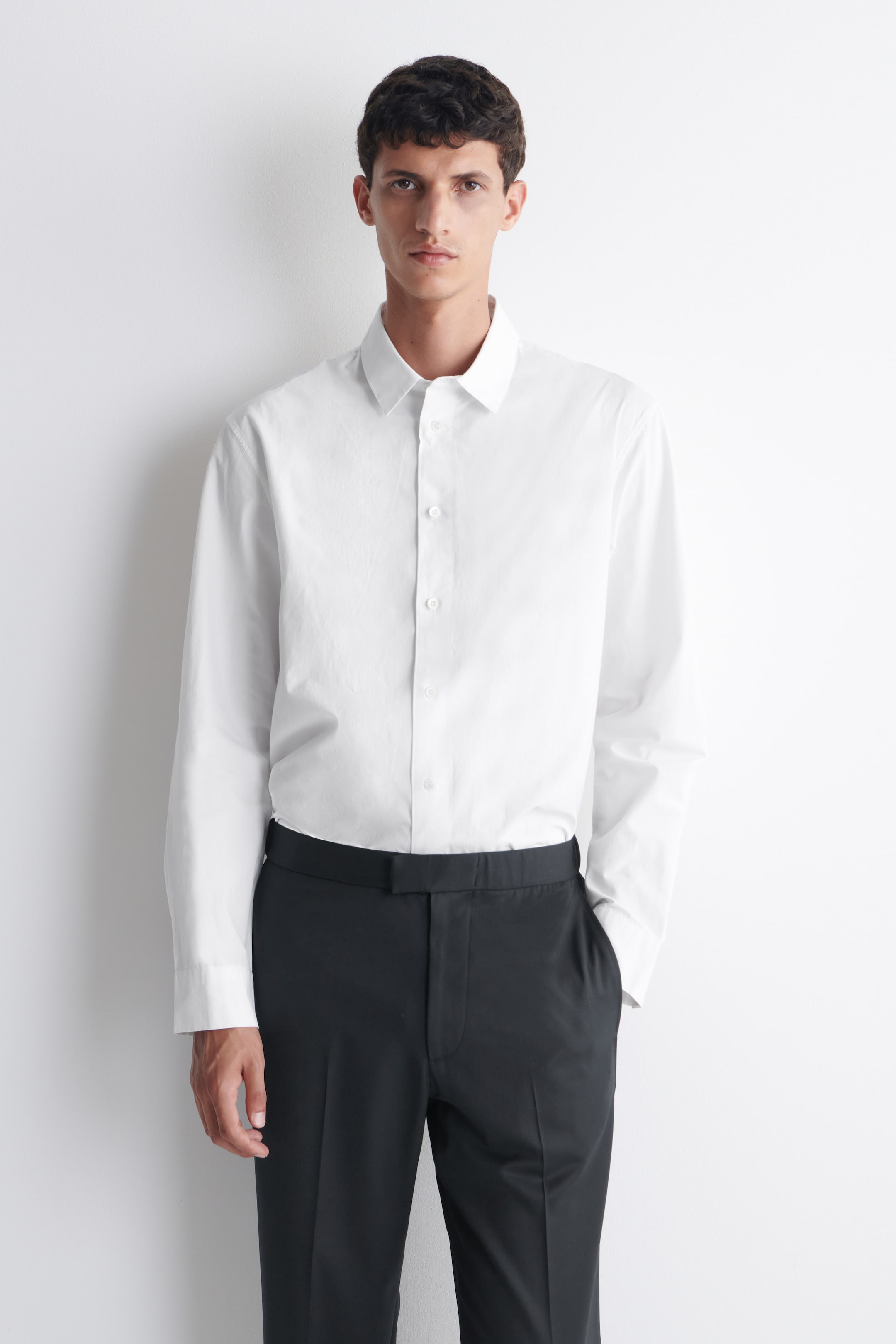 View larger image: The model is wearing a white long-sleeved shirt with a relaxed fit. The shirt boasts a classic collar and button-front fastening, with its long sleeves reaching the wrists, and is neatly tucked into dark trousers.