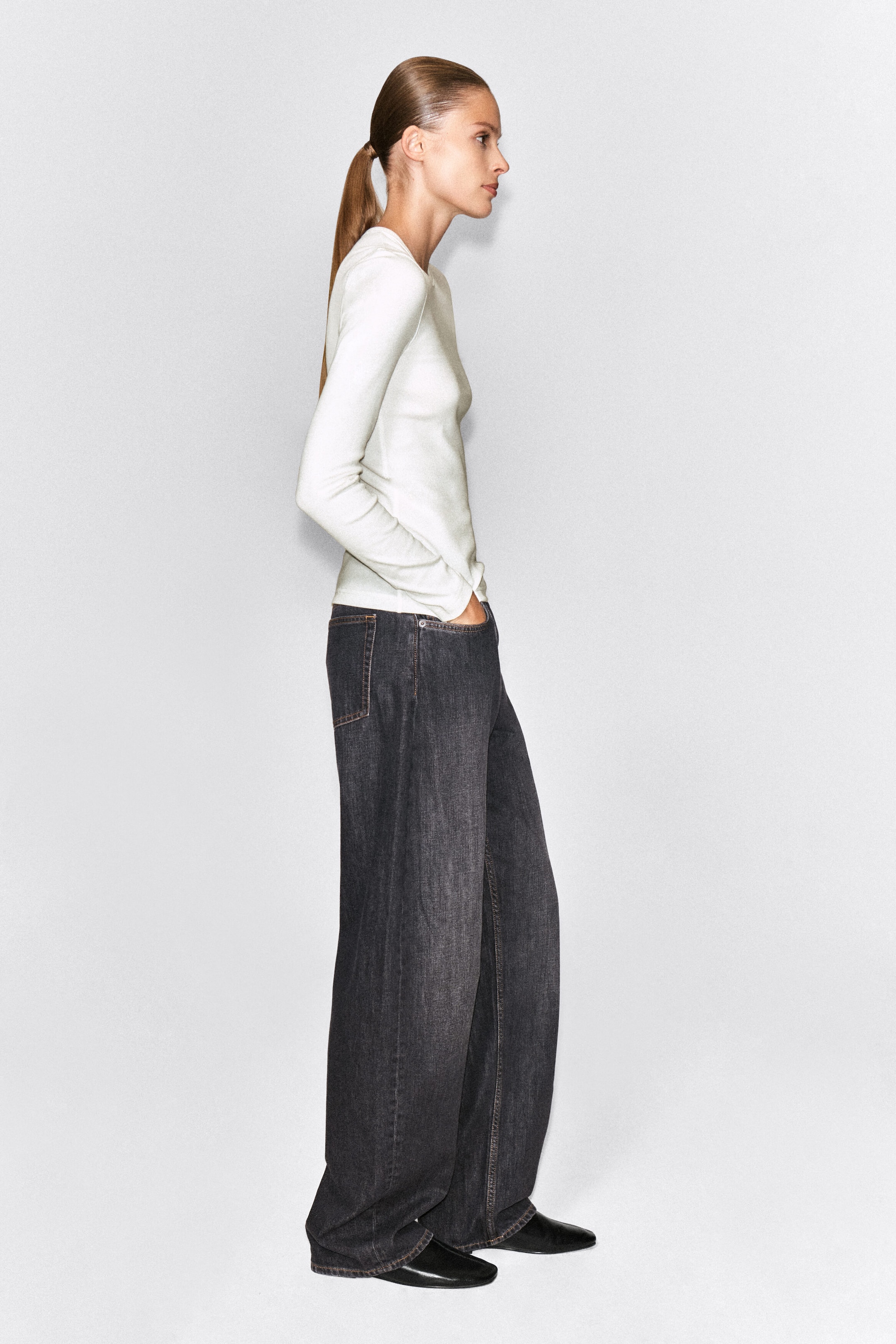 A slim, ribbed cotton long-sleeve top in off-white is worn by the model, who is standing in profile with her hands tucked into the pockets of dark, wide-leg jeans. The top features a crew neck and a fitted silhouette. A slim, ribbed cotton long-sleeve top in off-white is worn by the model, who is standing in profile with her hands tucked into the pockets of dark, wide-leg jeans. The top features a crew neck and a fitted silhouette.