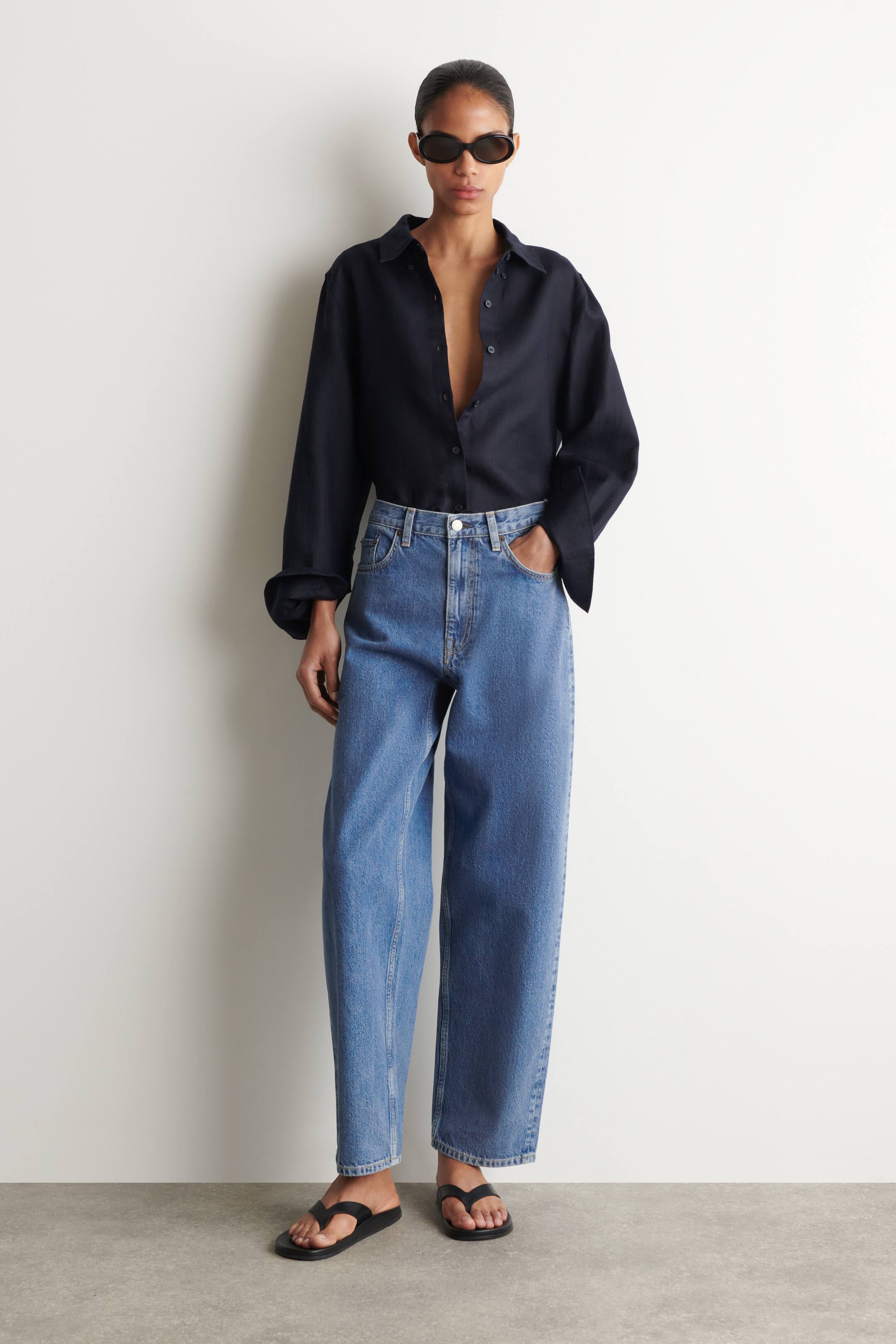Grösseres Bild ansehen: Ein dunkelblaues Langarmhemd wird getragen, teilweise aufgeknöpft und mit hochgekrempelten Manschetten, wobei es in hellblaue High-Waist-Jeans im Relaxed Fit gesteckt ist. Die Jeans zeigen einen sichtbaren vorderen Knopfverschluss und werden mit schwarzen Zehentrenner-Sandalen kombiniert.