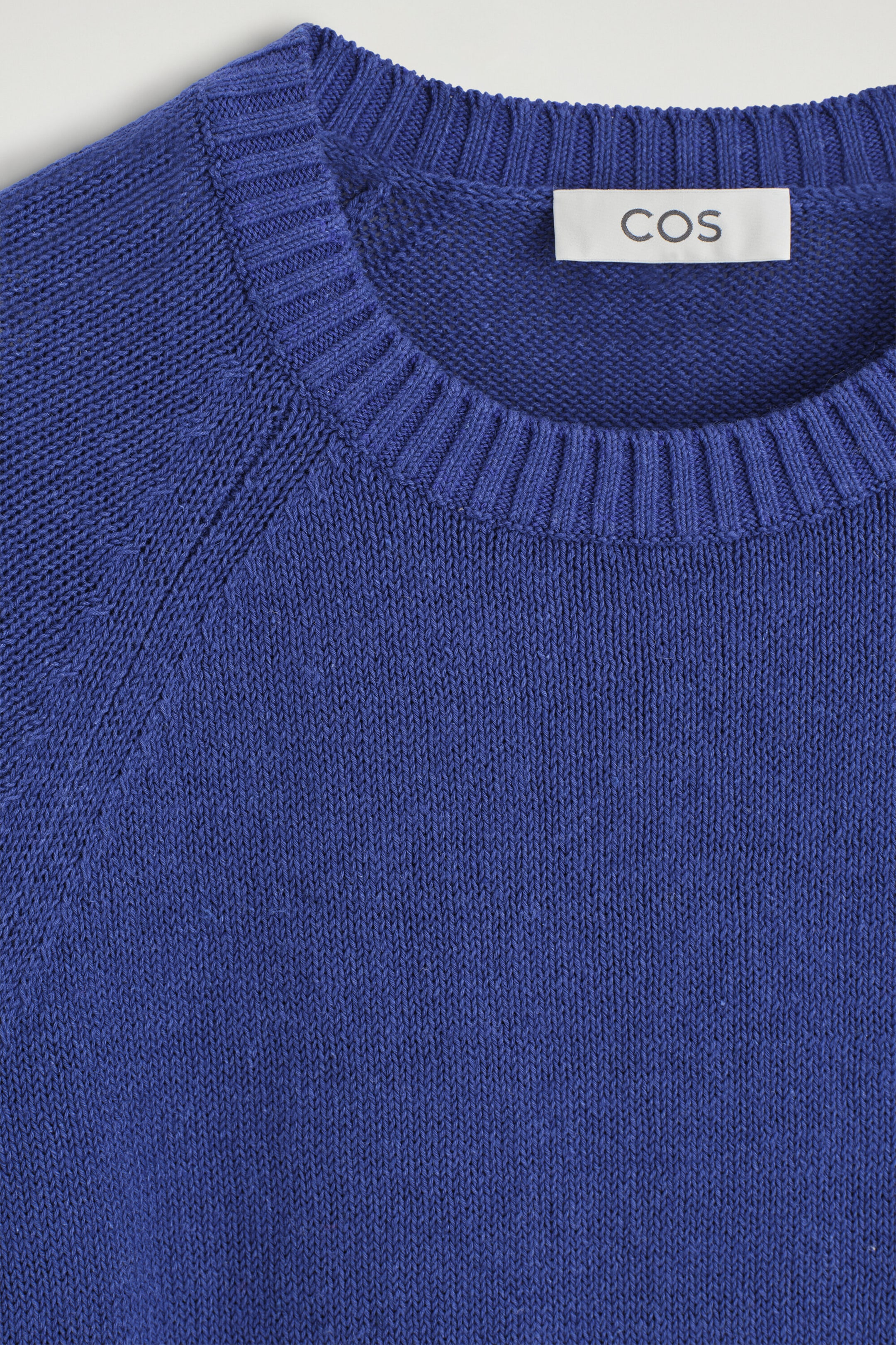 The Shrunken Knitted Linen T-Shirt showcases a close-up front view of its vibrant blue knit texture. It features a ribbed crew neckline, raglan sleeves, and a white brand label stitched inside the collar.