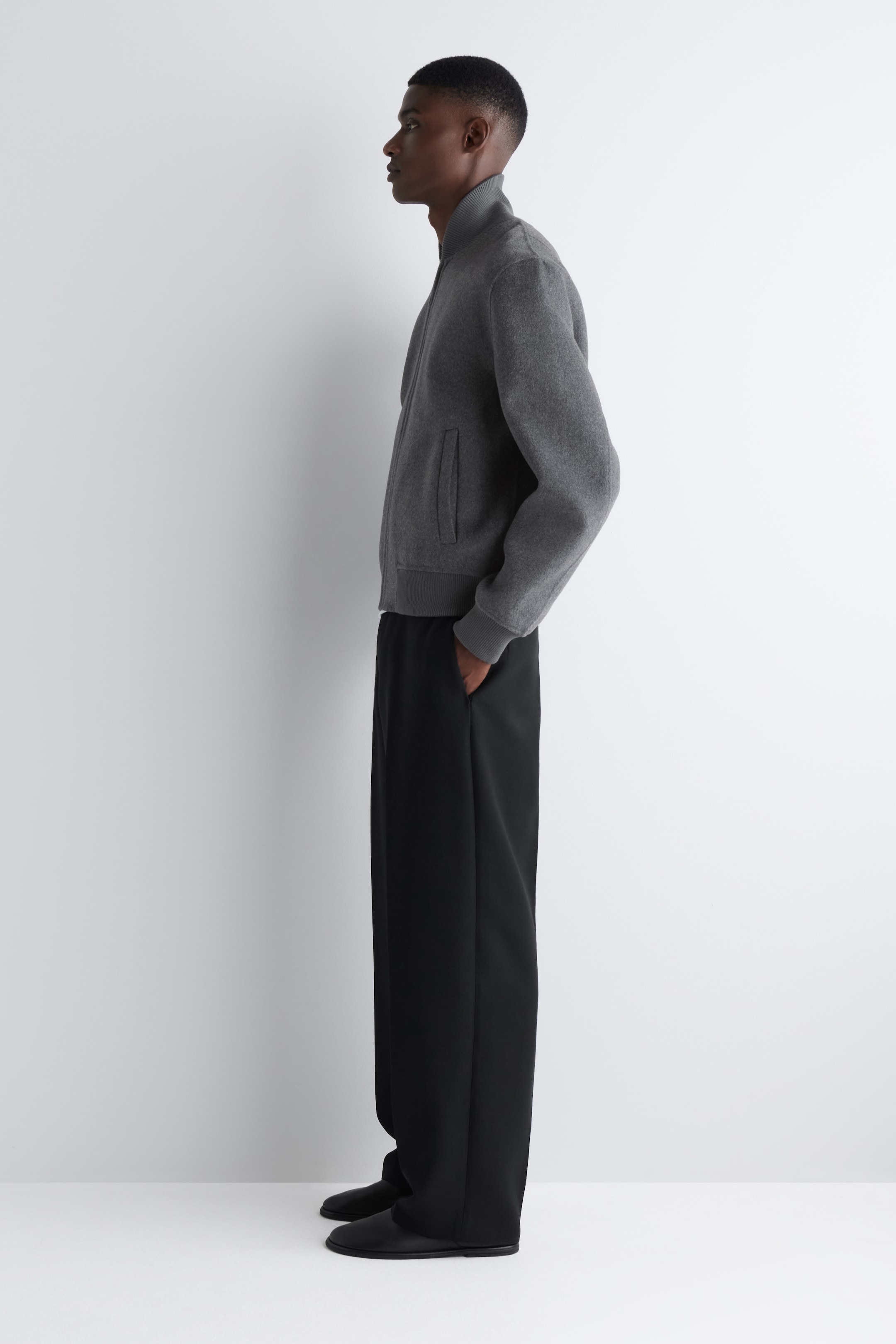 Ver imagen más grande: Una chaqueta bomber gris oscuro se muestra de perfil lateral, con un cuello alto acanalado, un cierre frontal completo, bolsillos de ojal y puños y dobladillo acanalados. La chaqueta se combina con pantalones de pierna ancha negros, que aparecen con un ajuste relajado y fluido.