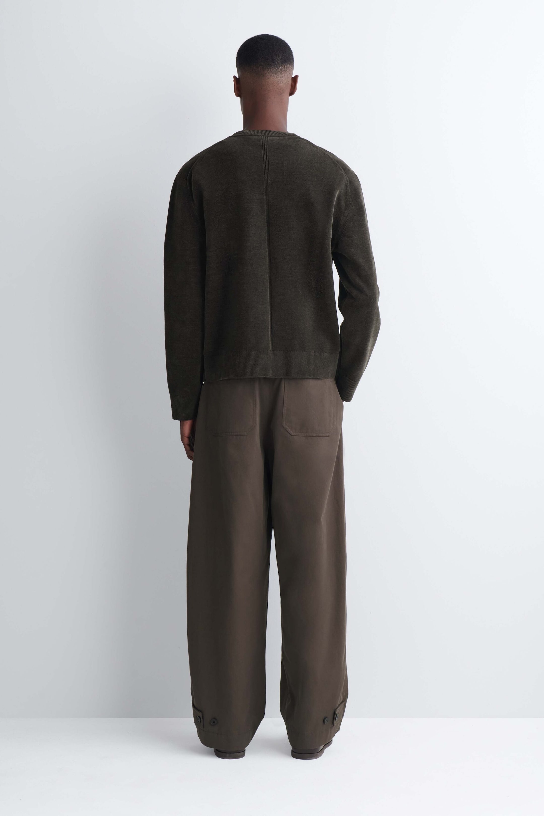 The model is wearing a dark olive-green cotton chenille crewneck sweater, facing away from the camera to showcase the garment’s textured finish and center back vertical seam. The sweater is styled with wide-leg brown trousers. The model is wearing a dark olive-green cotton chenille crewneck sweater, facing away from the camera to showcase the garment’s textured finish and center back vertical seam. The sweater is styled with wide-leg brown trousers.
