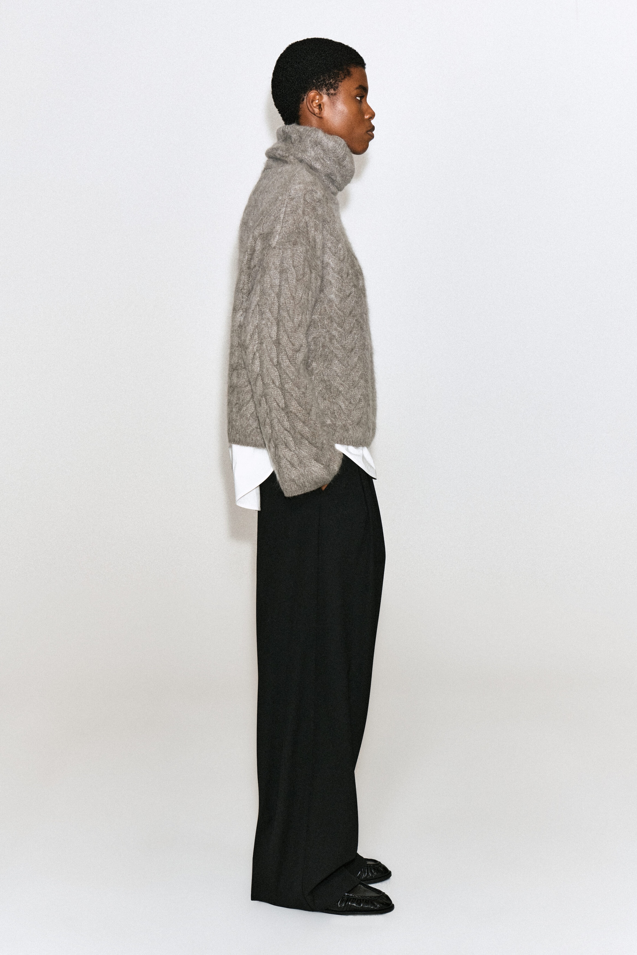 Un chandail col cheminée gris en cachemire brossé à maille torsadée est porté par le mannequin, qui se tient de profil. Le chandail texturé à col montant est superposé à une chemise blanche à col et est agencé avec un pantalon noir à jambes larges. Un chandail col cheminée gris en cachemire brossé à maille torsadée est porté par le mannequin, qui se tient de profil. Le chandail texturé à col montant est superposé à une chemise blanche à col et est agencé avec un pantalon noir à jambes larges.