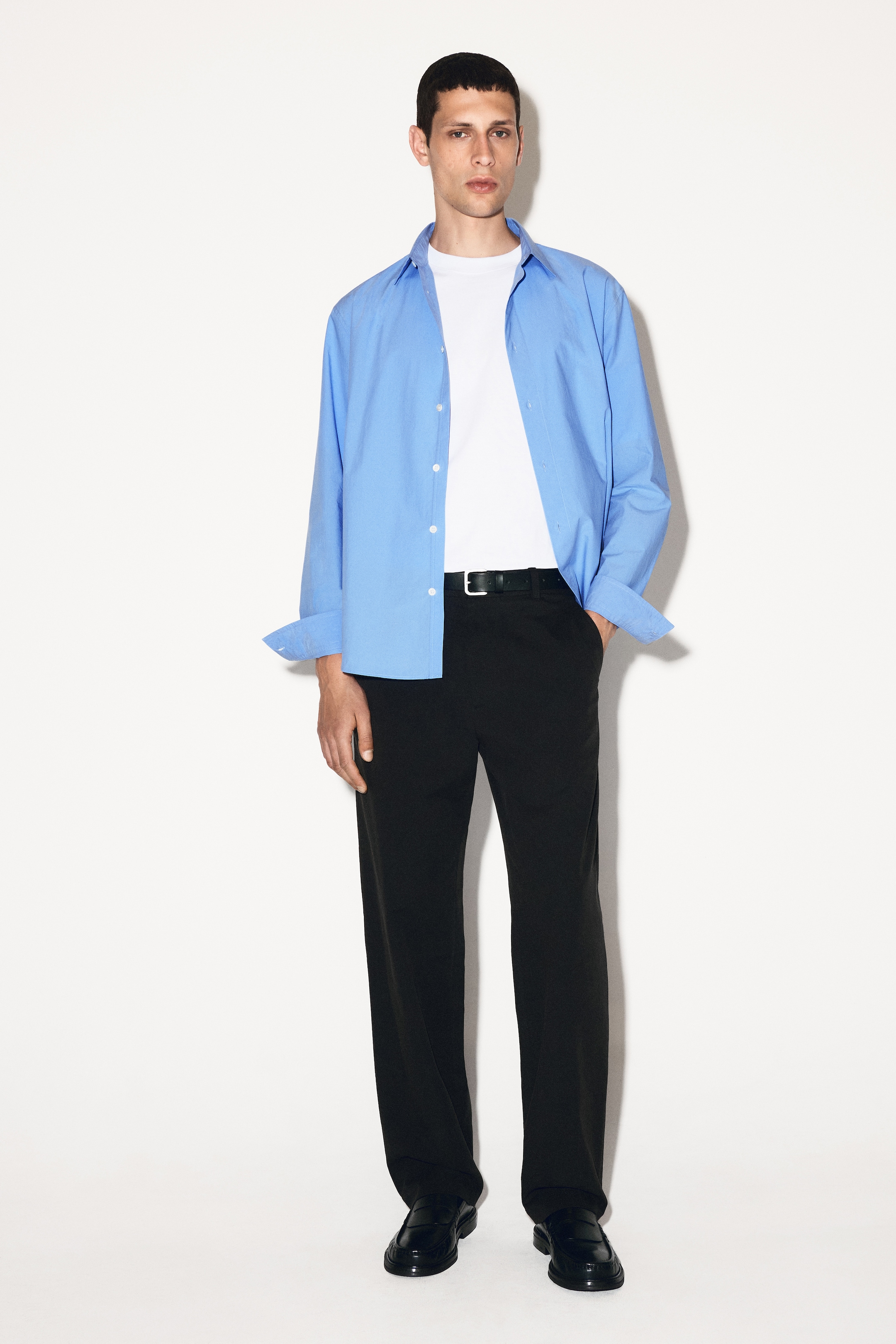 Agrandir l'image: Le produit est une chemise bleu ciel à manches longues, présentée de face avec une coupe décontractée. Ses boutons avant sont défaits, son col est ouvert et ses manches longues sont retroussées jusqu'aux avant-bras, avec un t-shirt blanc à col rond visible en dessous.