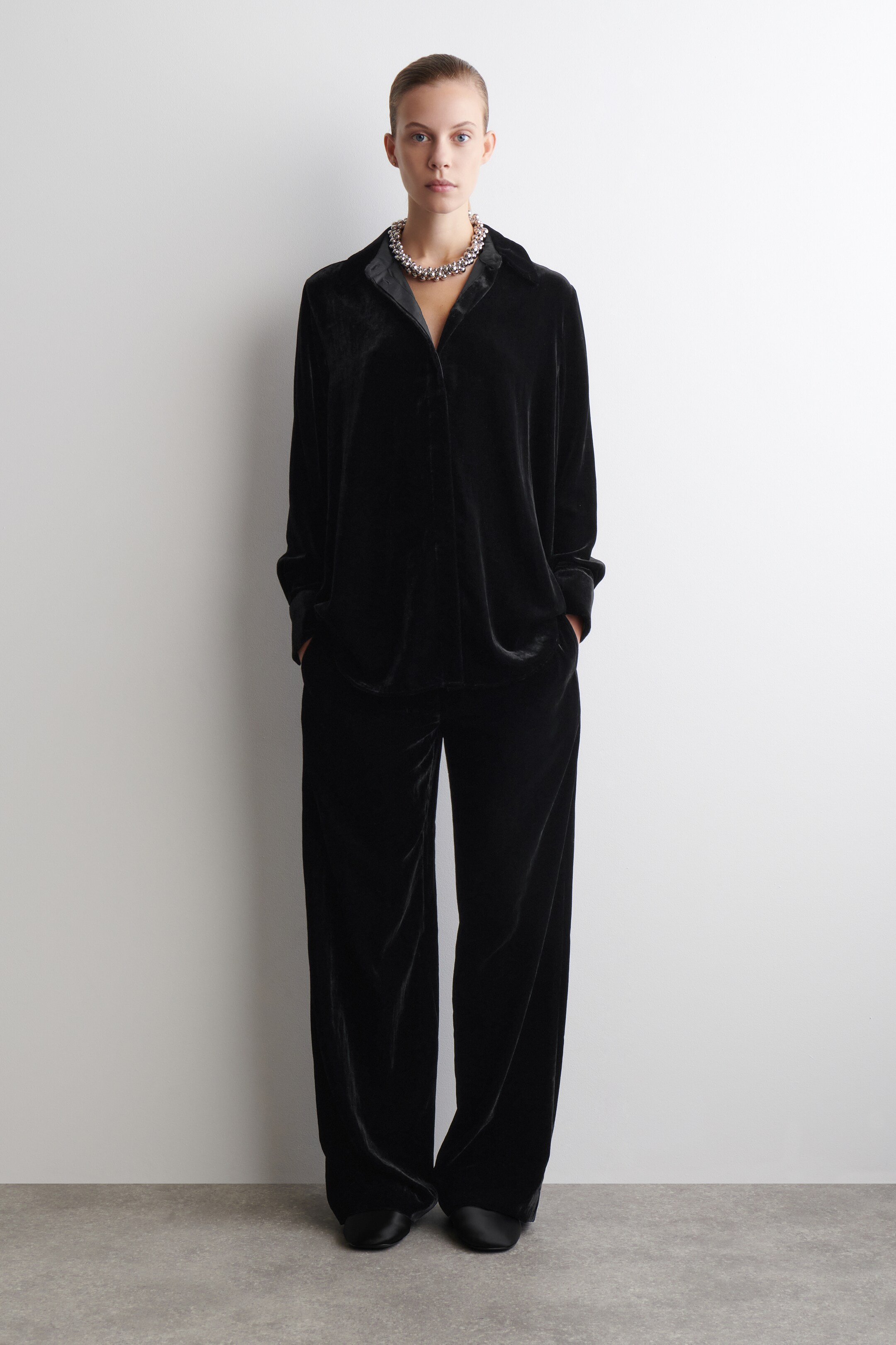 Visualizza immagine più grande: Un completo composto da una camicia a maniche lunghe di velluto nero e pantaloni a gamba larga è presentato, rivolto in avanti, con la camicia caratterizzata da un colletto a punta e i pantaloni che mostrano una vestibilità ampia. Una robusta collana color argento è indossata sopra la camicia, e scarpe nere piatte sono indossate con i pantaloni.