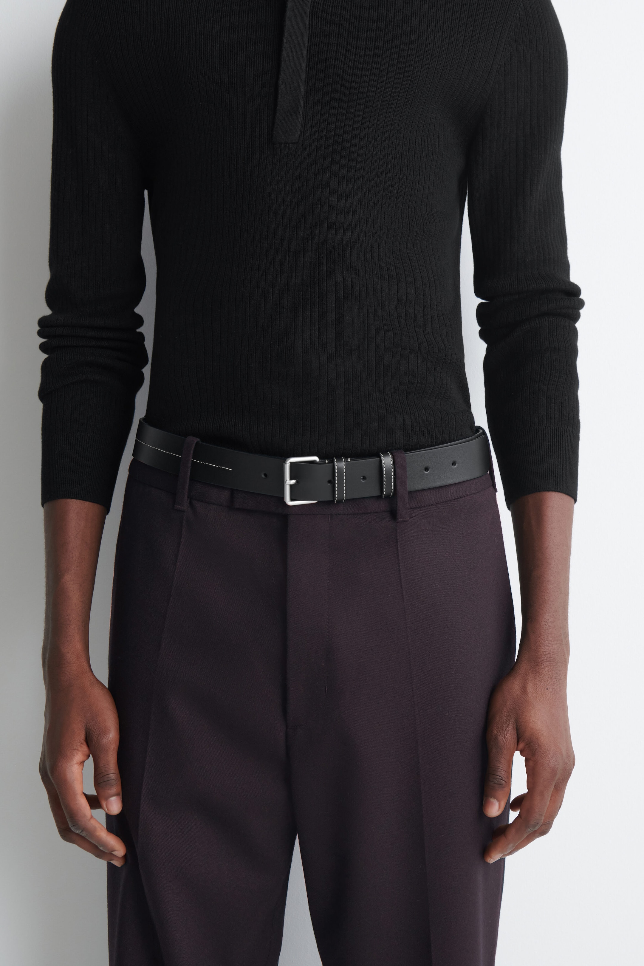 A black leather belt featuring white contrast stitching and a silver-toned metal buckle is being worn by the model. The belt is threaded through the belt loops of dark purple tailored trousers and paired with a black ribbed knit long-sleeve polo shirt. A black leather belt featuring white contrast stitching and a silver-toned metal buckle is being worn by the model. The belt is threaded through the belt loops of dark purple tailored trousers and paired with a black ribbed knit long-sleeve polo shirt.