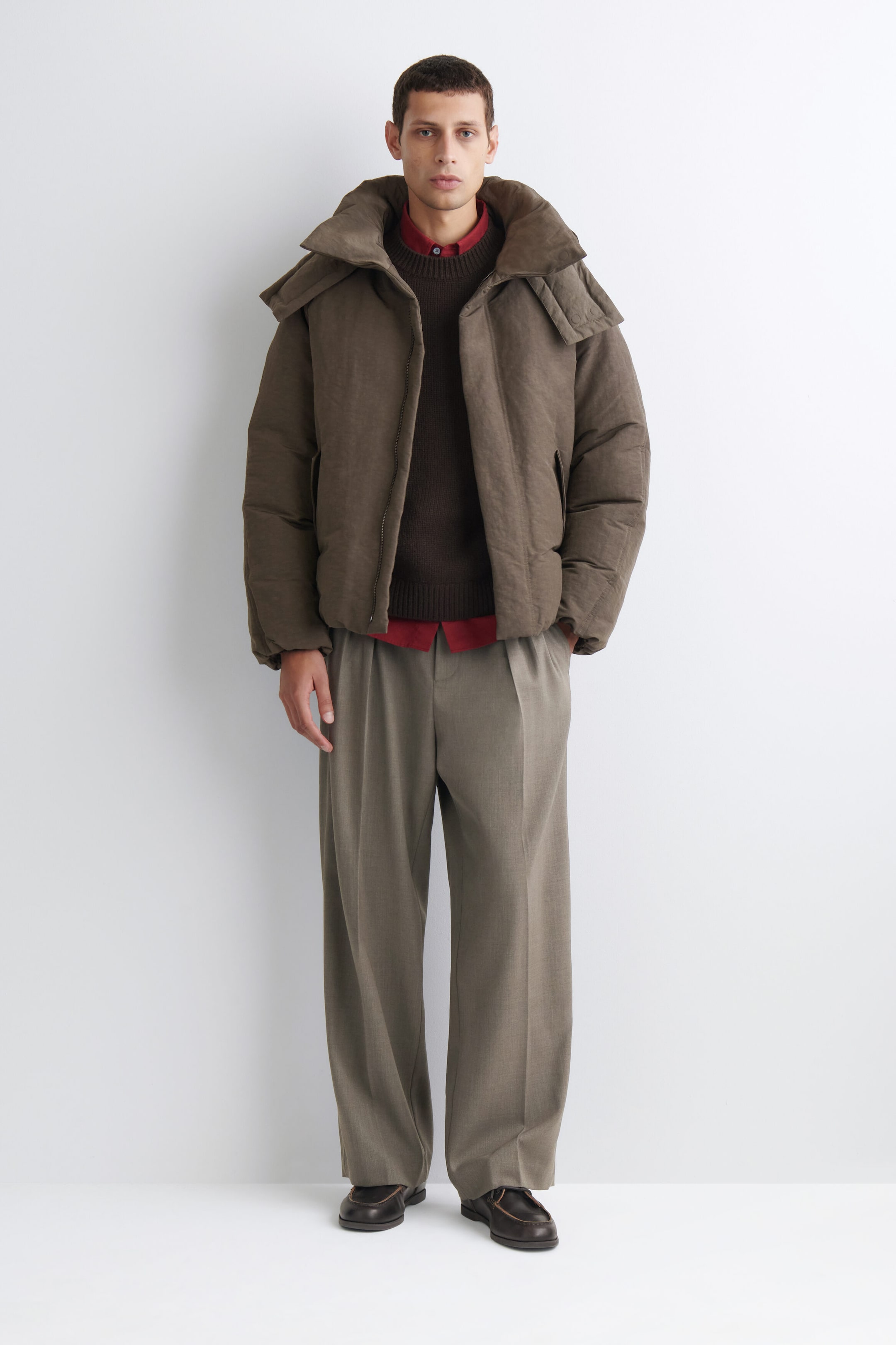 An oversized nylon-linen hooded down puffer jacket in textured brown is shown from the front, featuring a voluminous hood, a high collar, and a relaxed, puffy silhouette. The jacket is left open and layered over a dark brown knit sweater and a red collared shirt to emphasize its roomy fit. An oversized nylon-linen hooded down puffer jacket in textured brown is shown from the front, featuring a voluminous hood, a high collar, and a relaxed, puffy silhouette. The jacket is left open and layered over a dark brown knit sweater and a red collared shirt to emphasize its roomy fit.