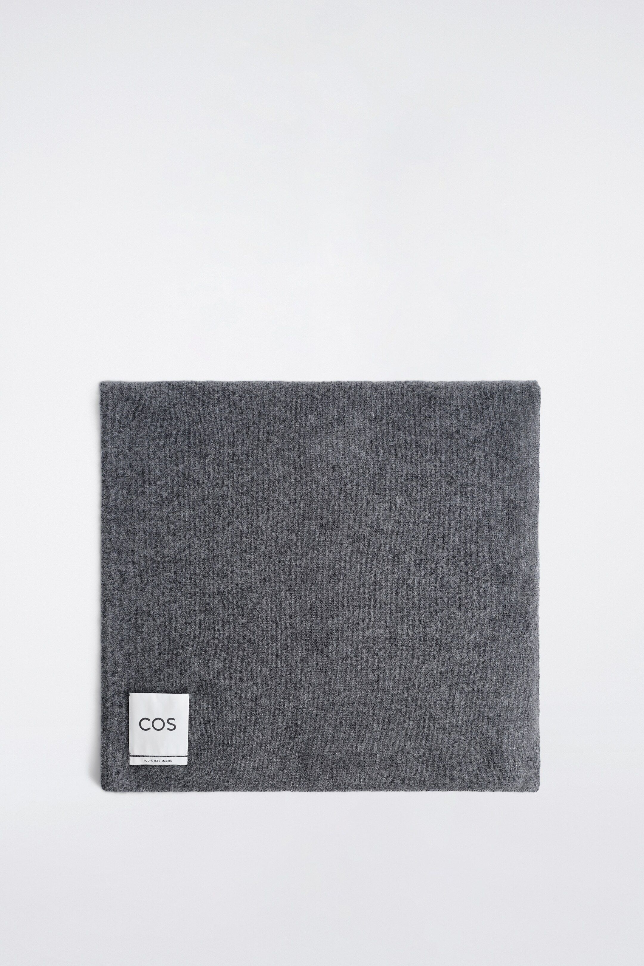 View larger image: A grey scarf is presented flat from a top view, displaying a finely textured surface. A white label with 'COS' and '100% CASHMERE' is visible on its bottom left corner.
