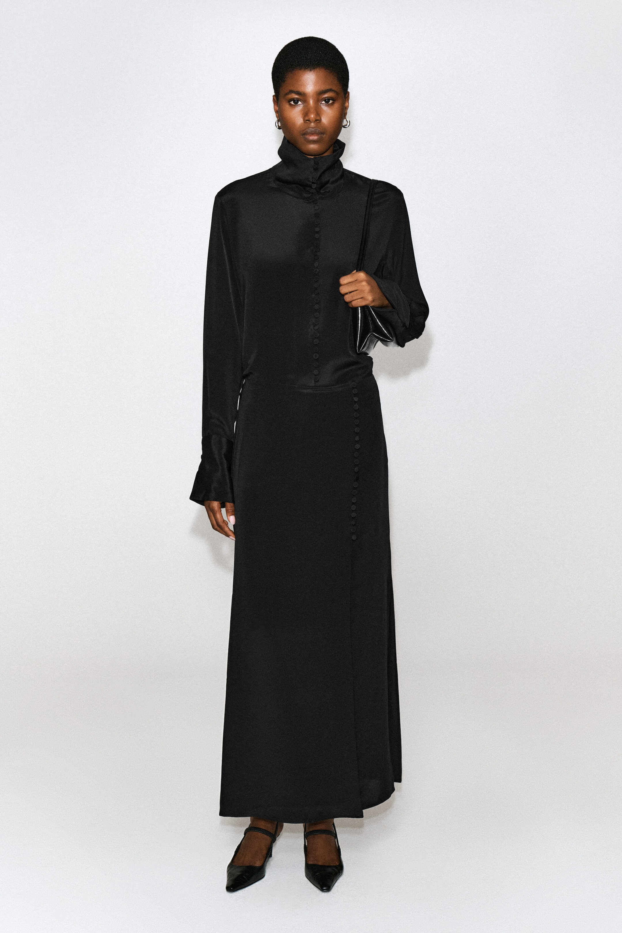A black long-sleeved maxi dress featuring a high funnel neck, a button-down front, and button detailing along the skirt is being worn by the model. The model is also carrying a small black leather shoulder bag and wearing black pointed-toe heels.