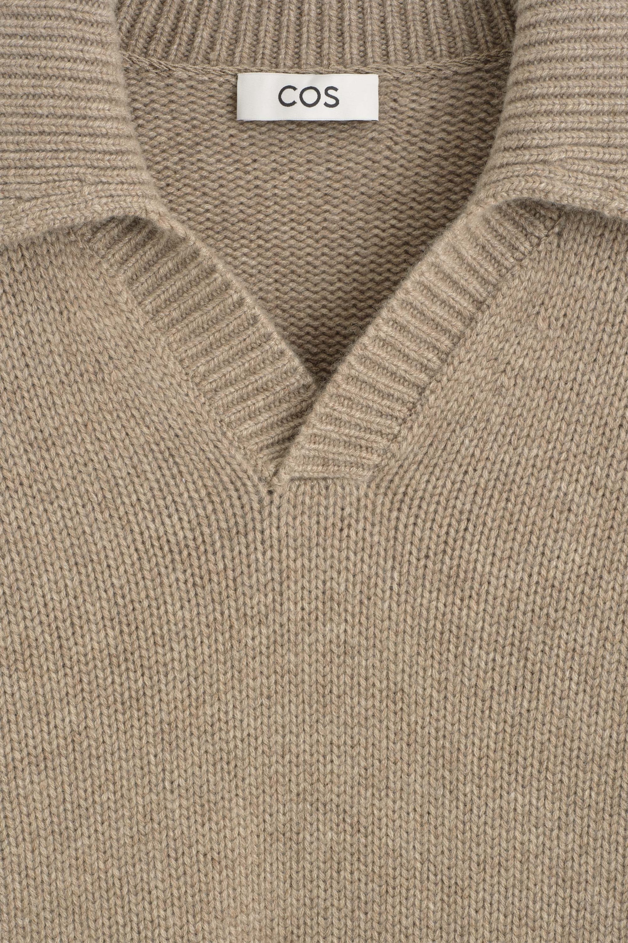 The chunky cashmere polo sweater is showcased in a close-up front view of the neckline, highlighting its textured beige knit. The design features a ribbed open polo collar and a V-neckline, with a white brand label visible on the inner collar. The chunky cashmere polo sweater is showcased in a close-up front view of the neckline, highlighting its textured beige knit. The design features a ribbed open polo collar and a V-neckline, with a white brand label visible on the inner collar.