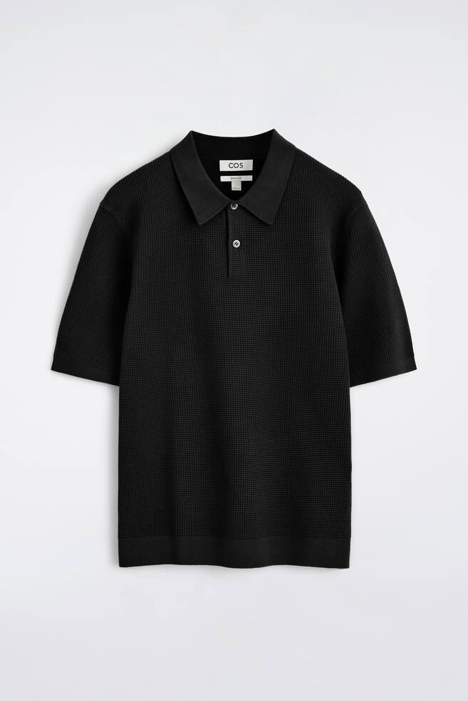 Ein schwarzes Polohemd wird von vorne präsentiert und verfügt über eine Mikro-Waffelstrickstruktur, einen gerippten Polokragen, eine Zweiknopfleiste mit hellen Knöpfen, kurze Ärmel mit gerippten Bündchen und einen gerippten Saum. - 1