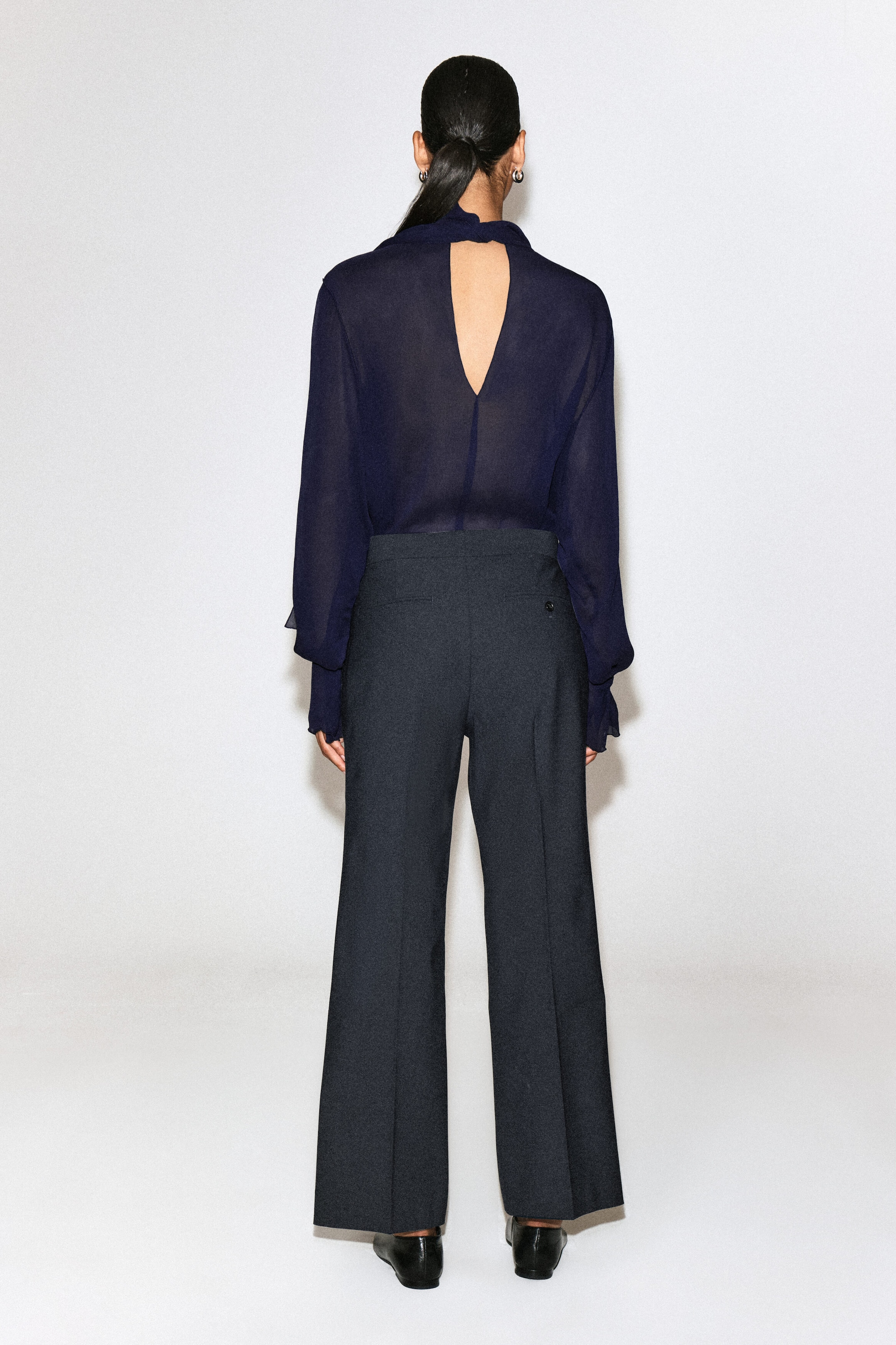 These twill cropped flared pants are shown from the back, featuring a high-rise waist, subtle rear welt pockets, and pressed creases running down the flared legs. The dark grey pants are styled with a tucked-in navy blue blouse to highlight their structured fit and clean waistband.