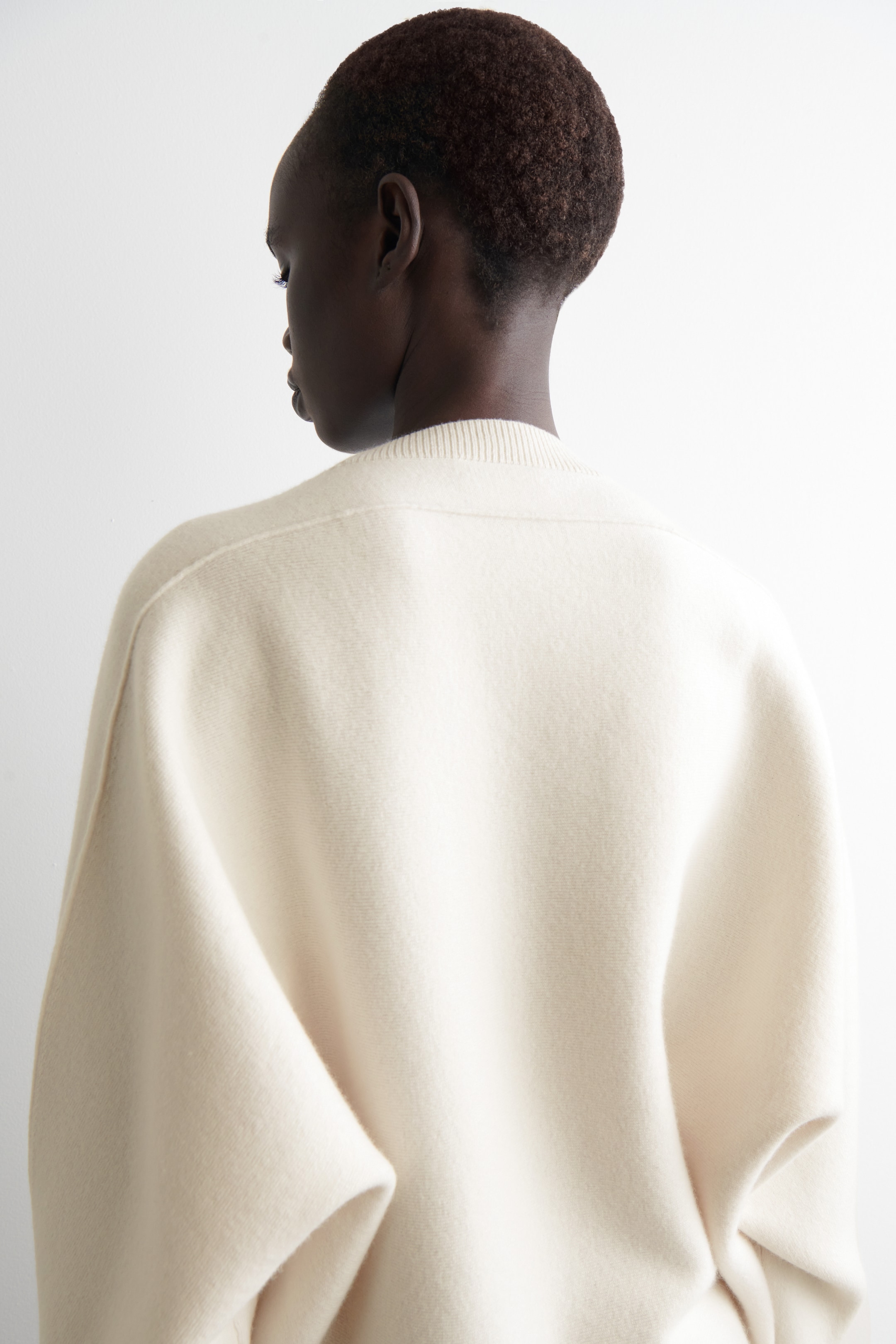 Visualizza immagine più grande: Un capo in maglia color crema è visto da dietro, mostrando la sua vestibilità rilassata e oversize. Presenta un collo a girocollo a coste e cuciture delle spalle abbassate, con maniche voluminose che si drappeggiano liberamente dalle spalle.