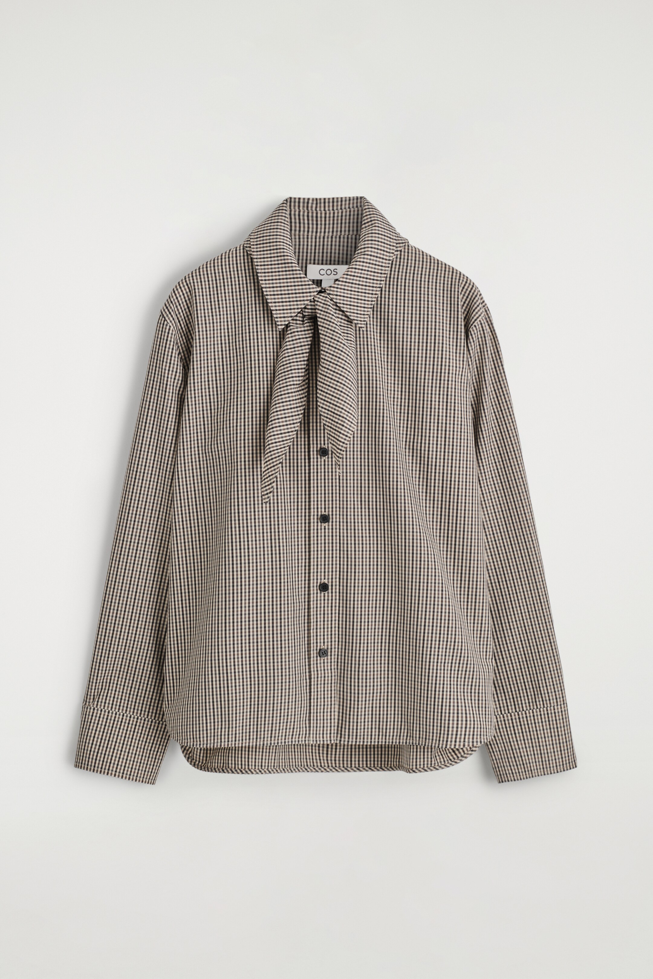 Ver imagem maior: VISTA FRONTAL. Uma camisa de micro-xadrez em castanho, preto e creme apresenta uma gola pontiaguda com um detalhe de gravata integrada atada na frente. A camisa é desenhada com mangas compridas, punhos largos e um fecho frontal com botões escuros.