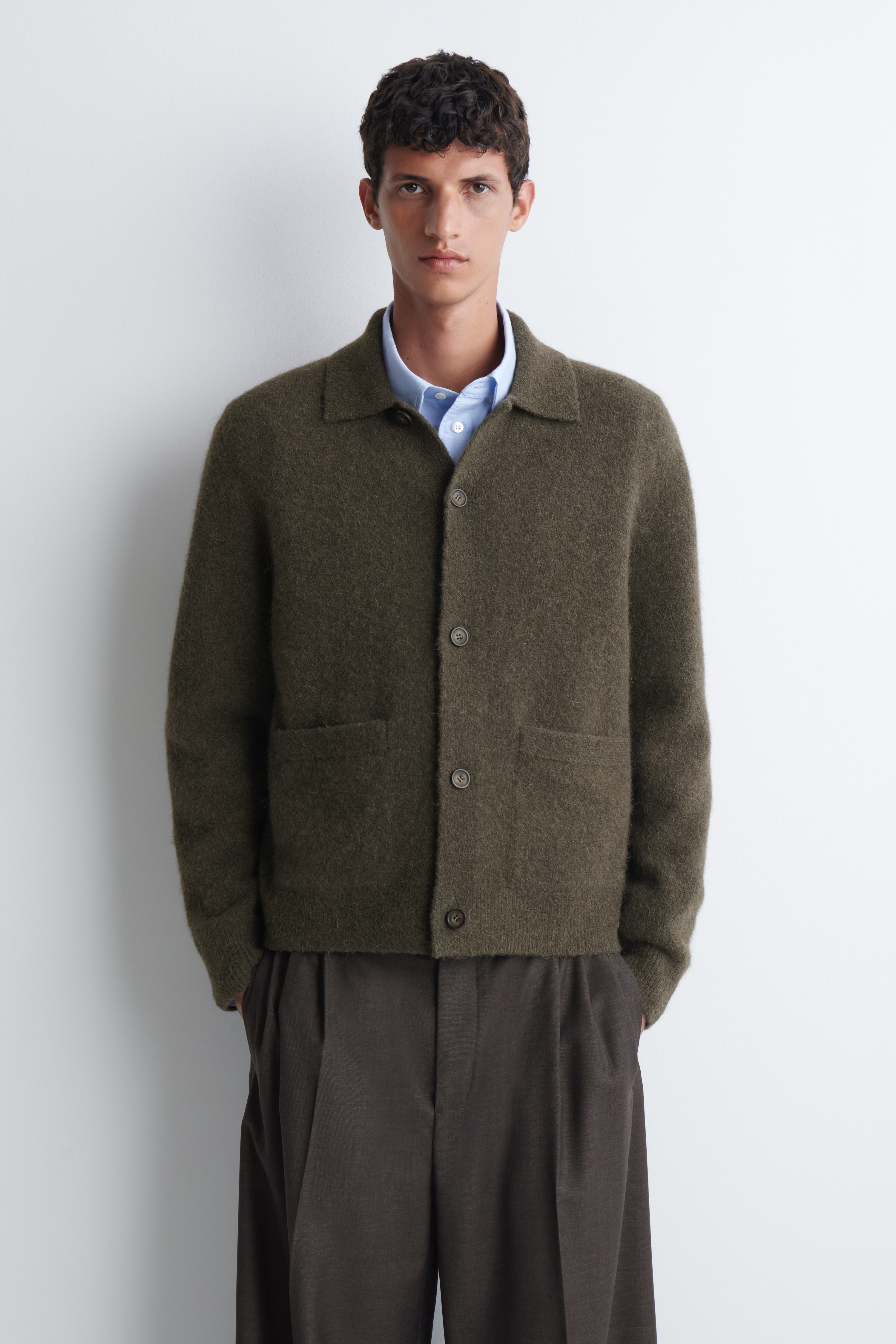 A textured collared cardigan is facing front, showcasing a soft, fuzzy knit in a dark olive green hue. It features a spread collar, a five-button front fastening, two front welt pockets, and ribbed cuffs.